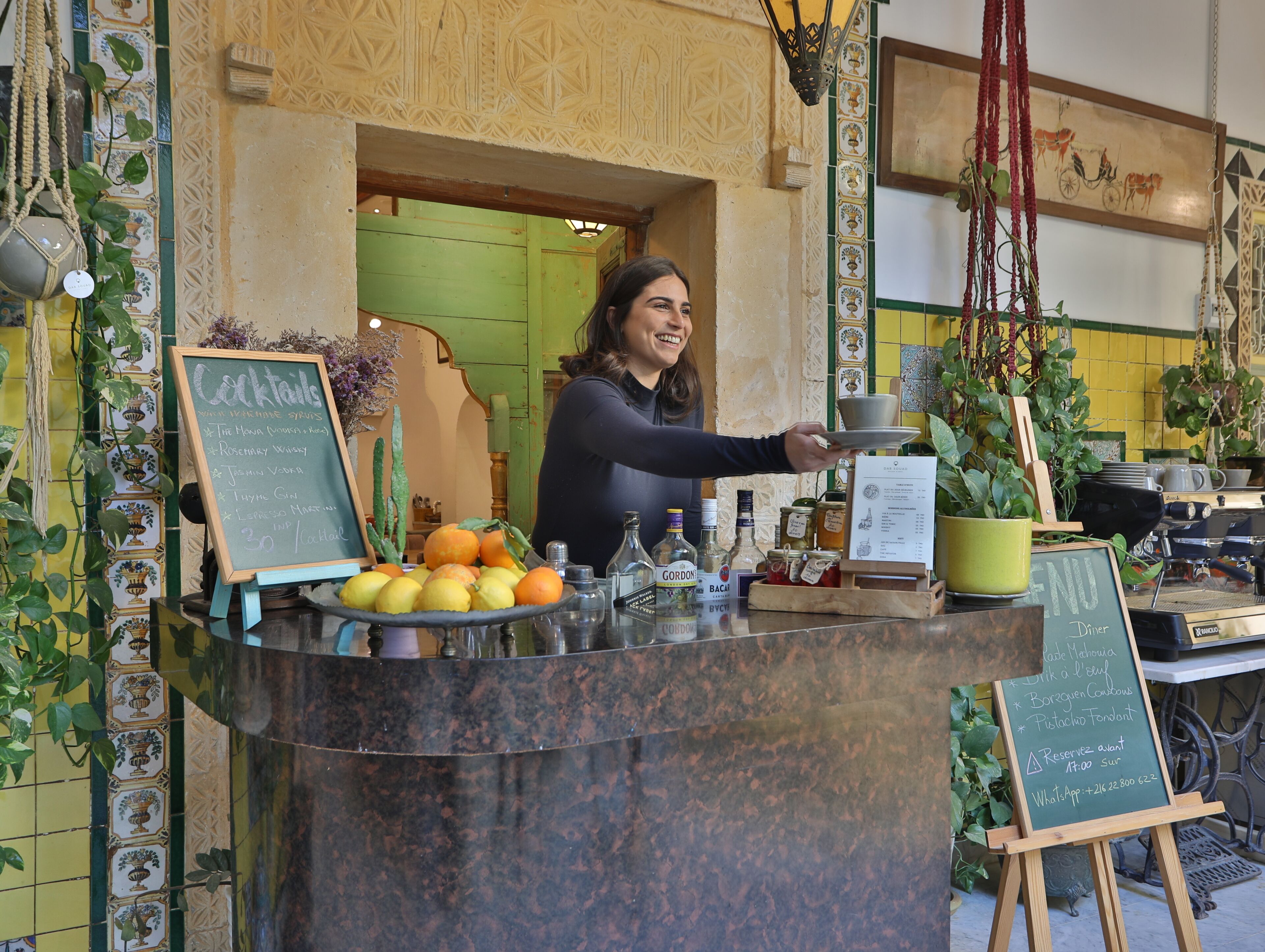 A bar counter with a chalkboard menu listing cocktails and prices, a variety of bottles, and a potted plant.