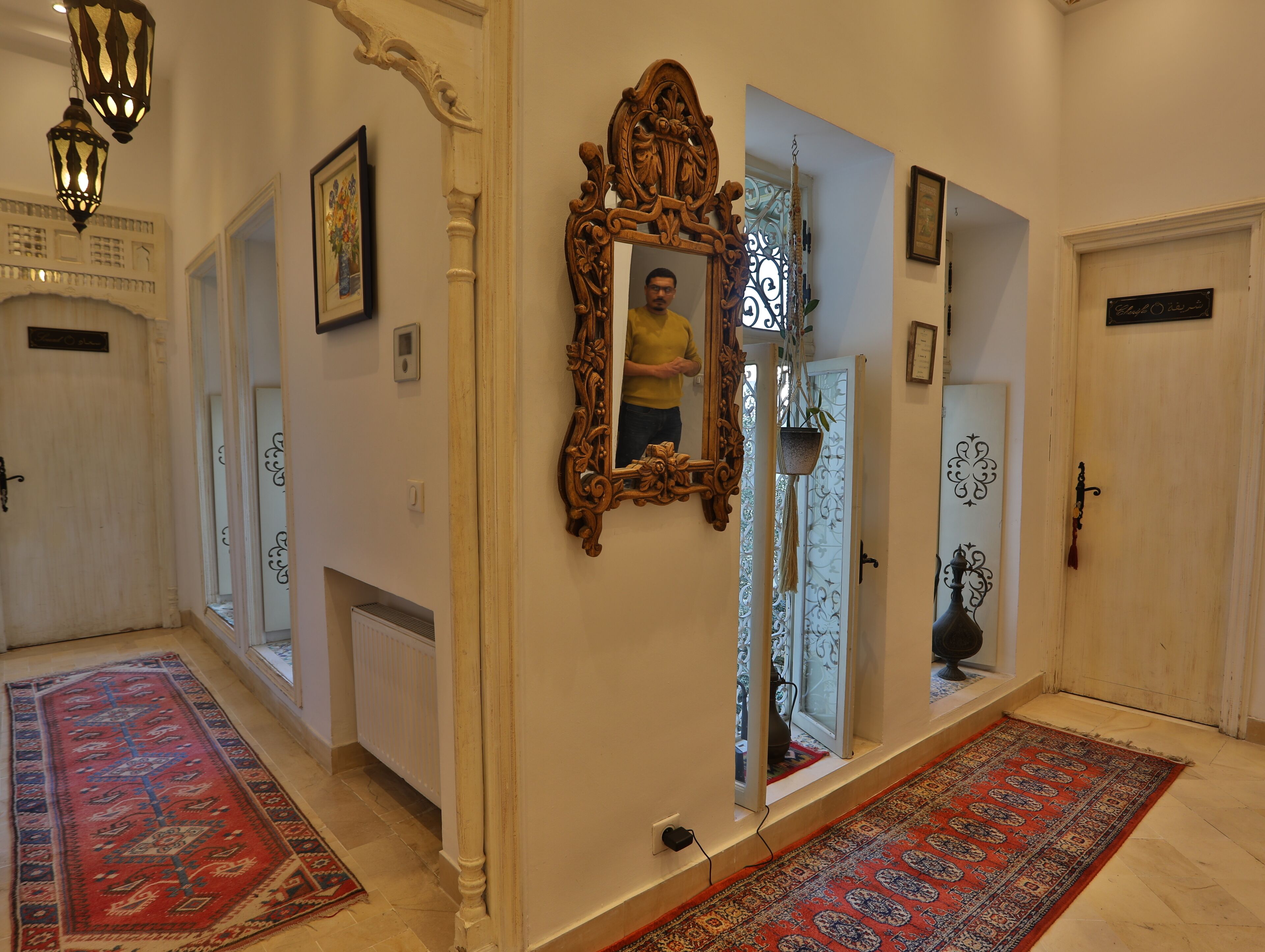 Un pasillo con puertas de madera ornamentadas, un espejo grande y alfombras estampadas.