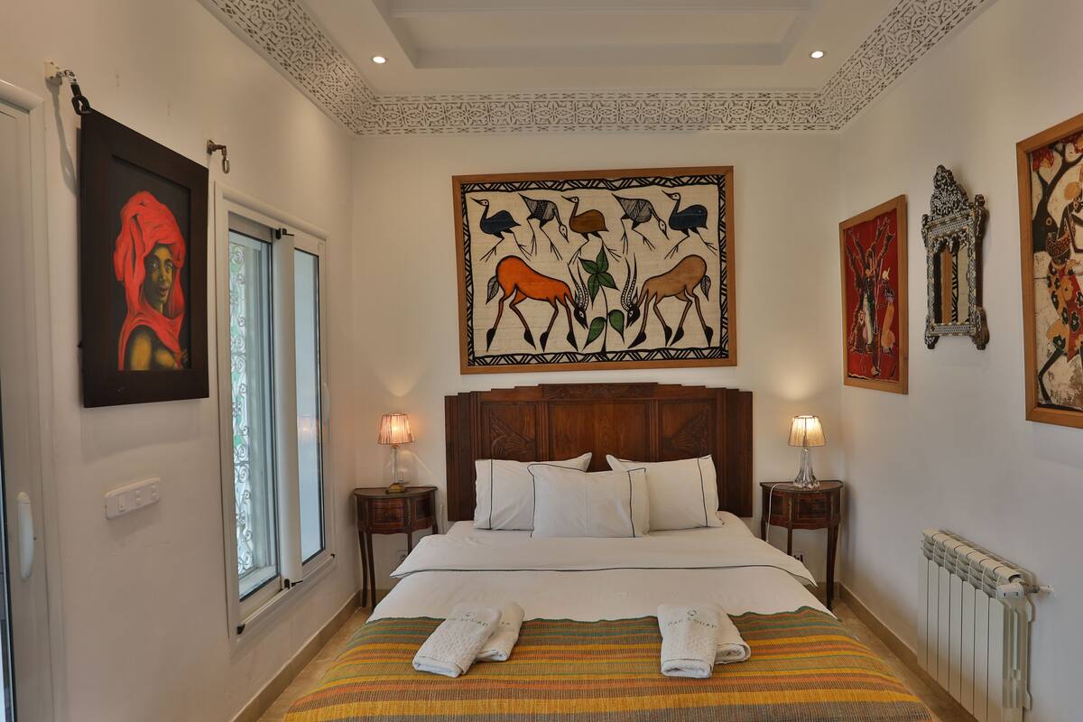 Una habitación de hotel con una cama grande, dos mesitas de noche con lámparas, una ventana con cortinas y varias obras de arte enmarcadas en las paredes.