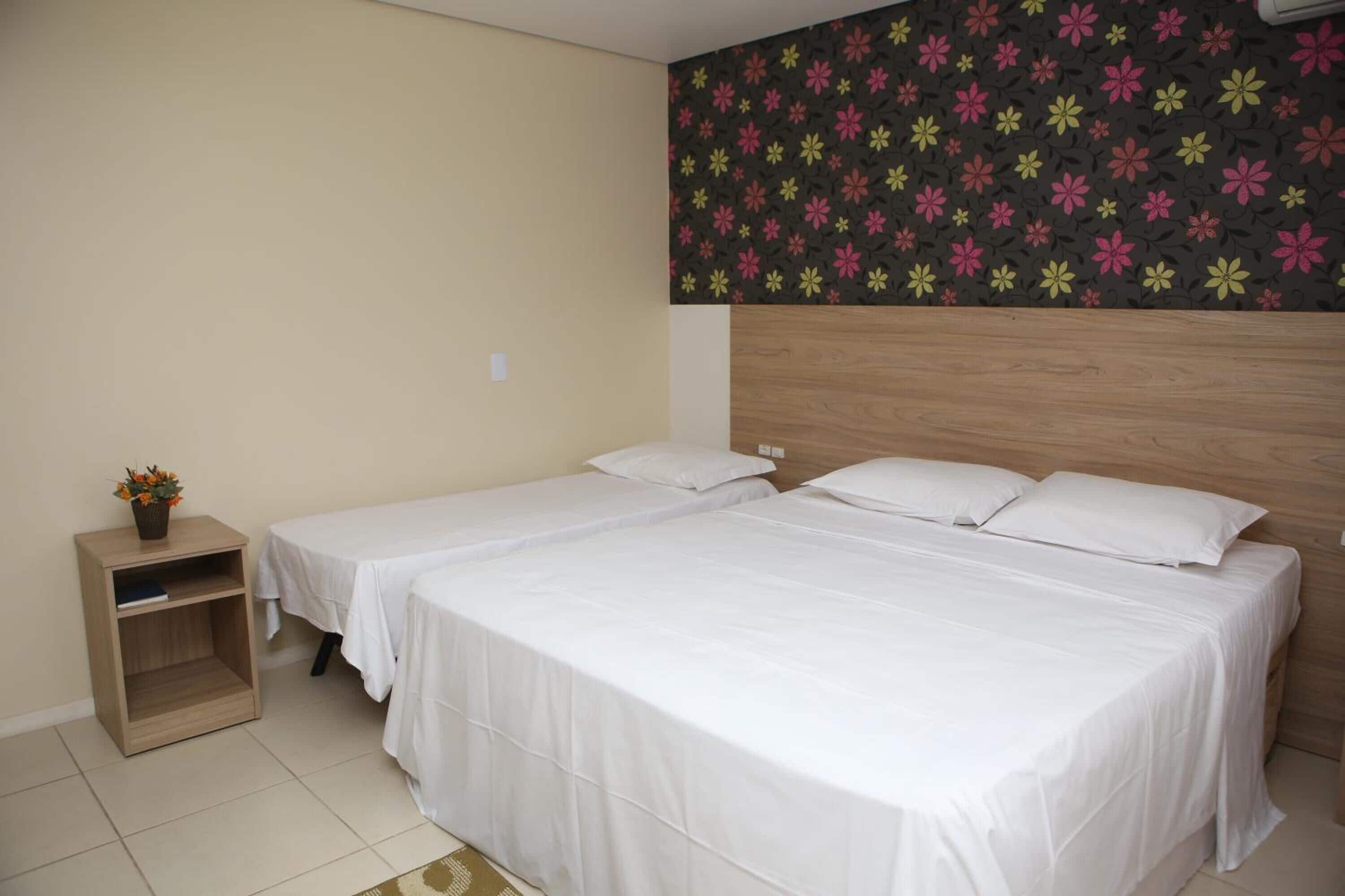 Une chambre d’hôtel avec deux lits simples, une table de chevet en bois ornée d’un vase de fleurs et une tête de lit à motifs floraux.