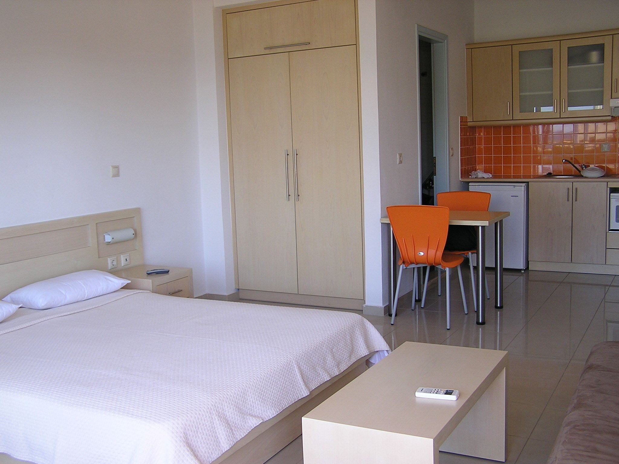A hotel room with a bed, a desk, an orange chair, and a kitchenette.