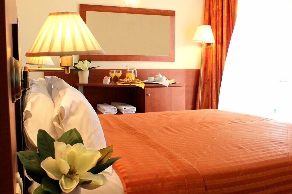 Habitación de hotel con cama, mesita de noche con lámpara, espejo, pequeña mesa con provisiones para el desayuno y ventana con cortinas.