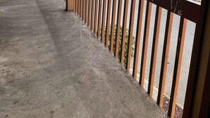 Un espacio al aire libre techado, con piso de concreto y barandilla de metal, que da vista a una calle y áreas verdes.