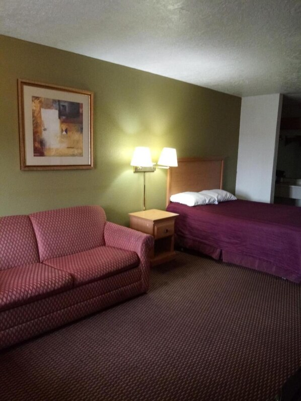 Habitación de hotel con un sofá rojo estampado, una mesita de noche de madera con dos lámparas, una cama con colcha púrpura y un cuadro enmarcado en la pared.