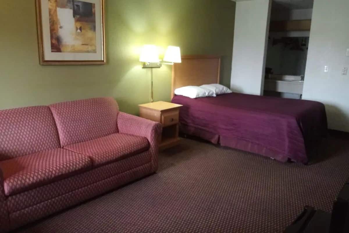 Quarto de hotel com um sofá vermelho estampado, uma cama com colcha roxa, uma mesinha de cabeceira com duas luminárias e um quadro na parede.