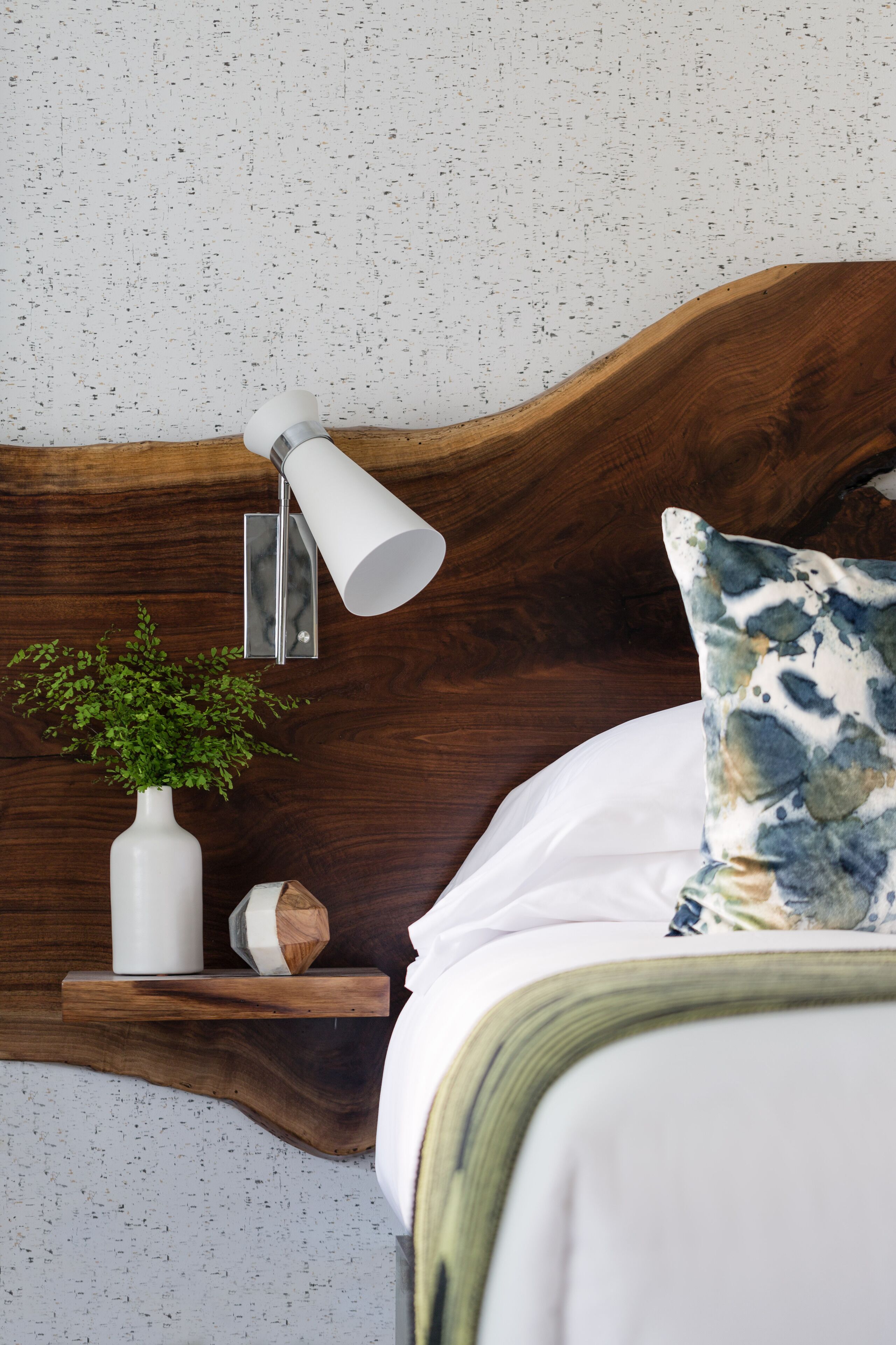 A bedroom with a wooden headboard, a bedside table with a white vase and a plant, a white lamp, and a floral pillow.