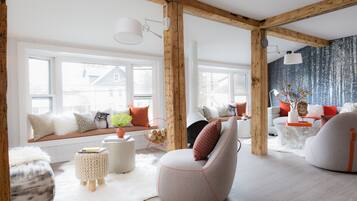 A modern living room with a large window, wooden beams, and a cozy seating area with cushions and a fireplace.