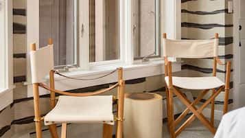 Una habitación con dos sillones de director de madera, una mesita redonda y una ventana que da a una pared con estampado.
