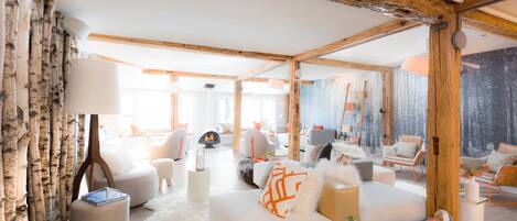 Un ampio soggiorno con soffitto alto, travi in legno e una grande finestra. Sono presenti diverse zone relax, tra cui un divano e poltrone, e un caminetto.