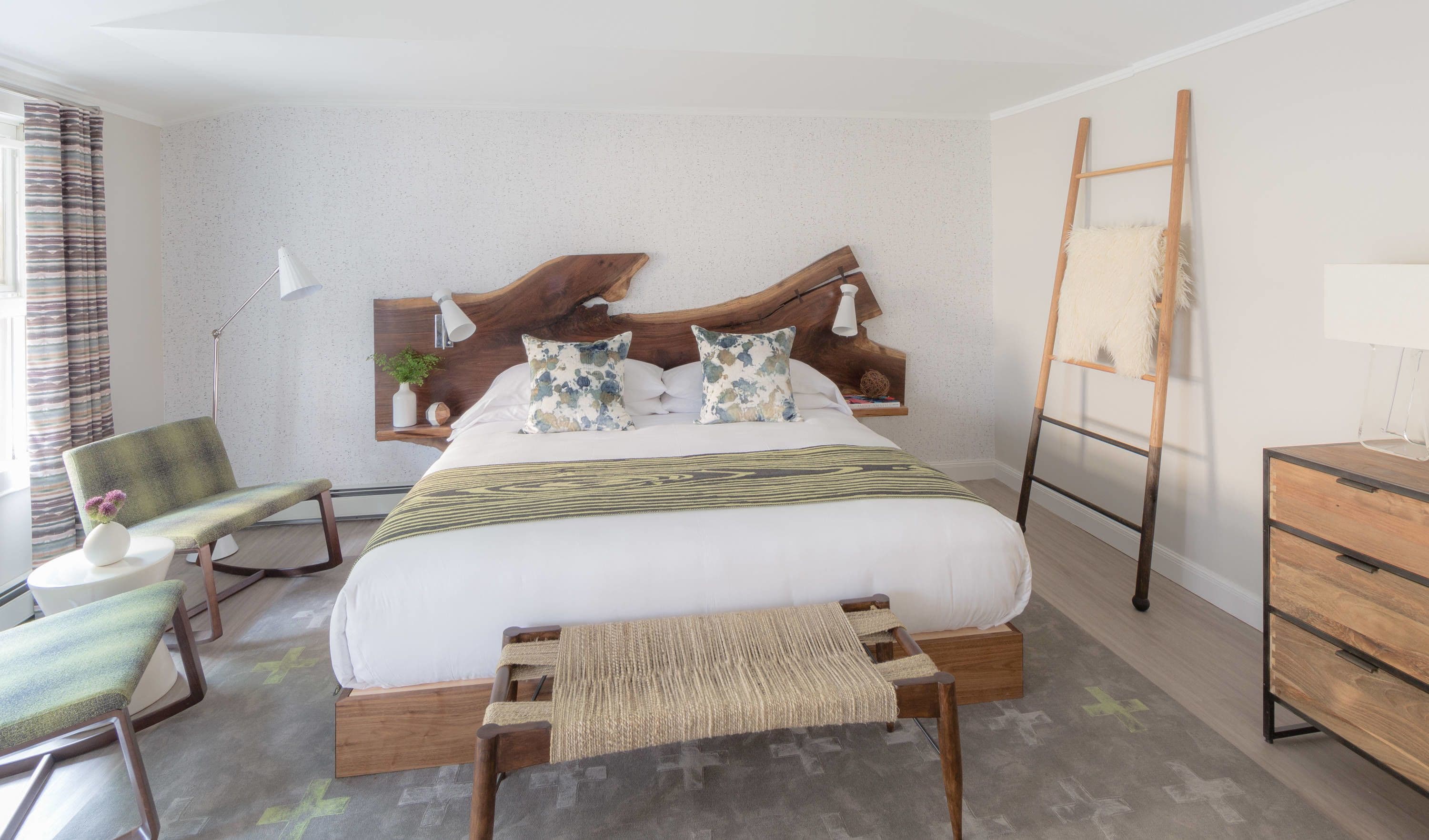 A neatly arranged bedroom with a large bed, a wooden headboard, a bedside table with a lamp, a chair, a small table with a vase, and a ladder against the wall.