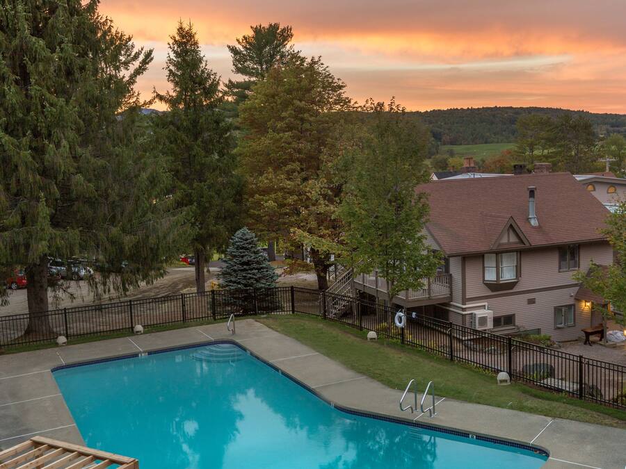 A rectangular swimming pool with a wooden deck and lounge chairs, surrounded by a fenced yard with trees and a house in the background.