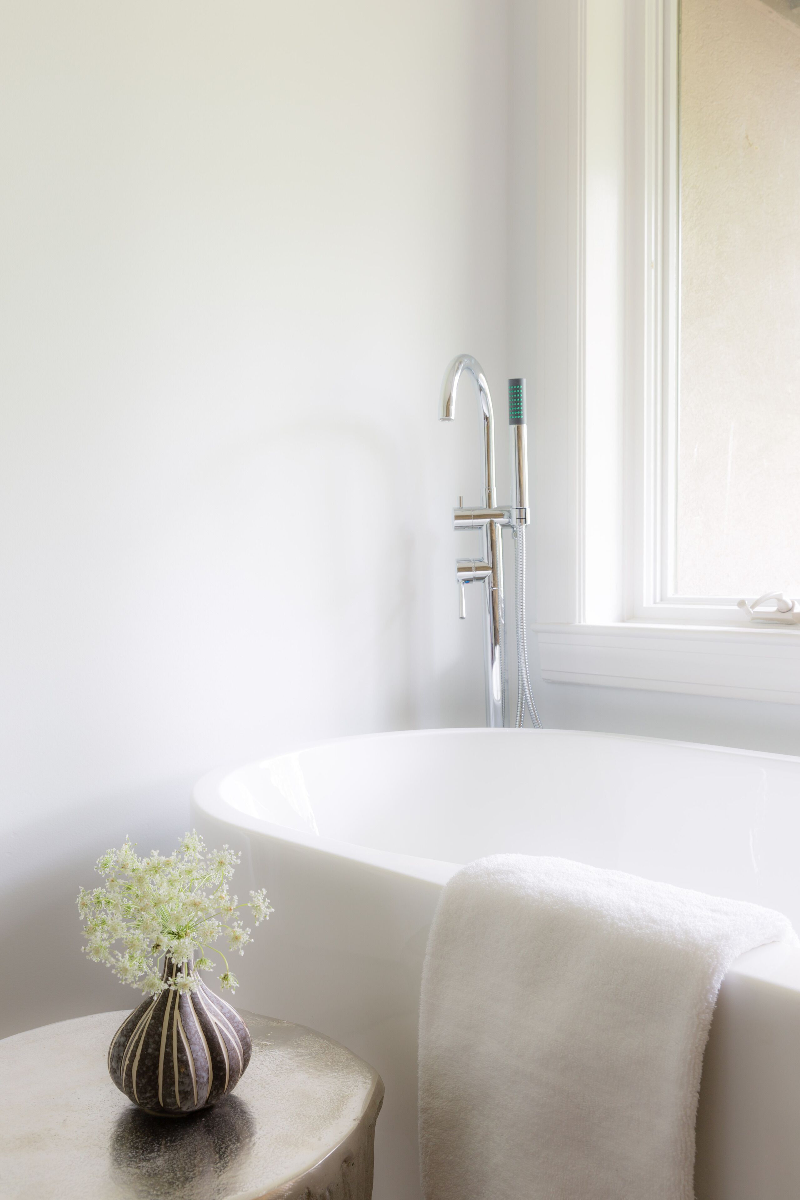 Un baño con una bañera blanca, cabezal de ducha y un florero con flores sobre una mesita.