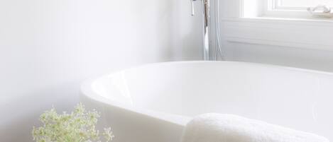 Un baño con una bañera blanca, cabezal de ducha y un florero con flores sobre una mesita.