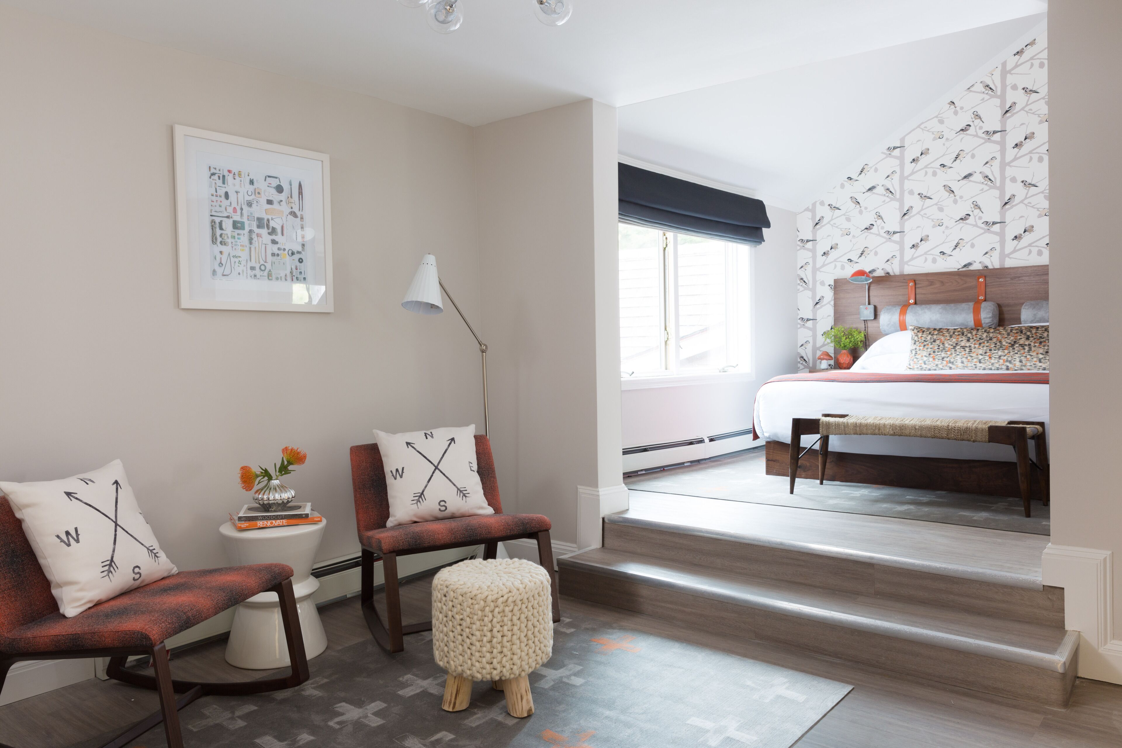 Un dormitorio moderno con una cama grande, una zona de estar pequeña con sillas, una mesita de noche con lámpara y una ventana con vistas al exterior.