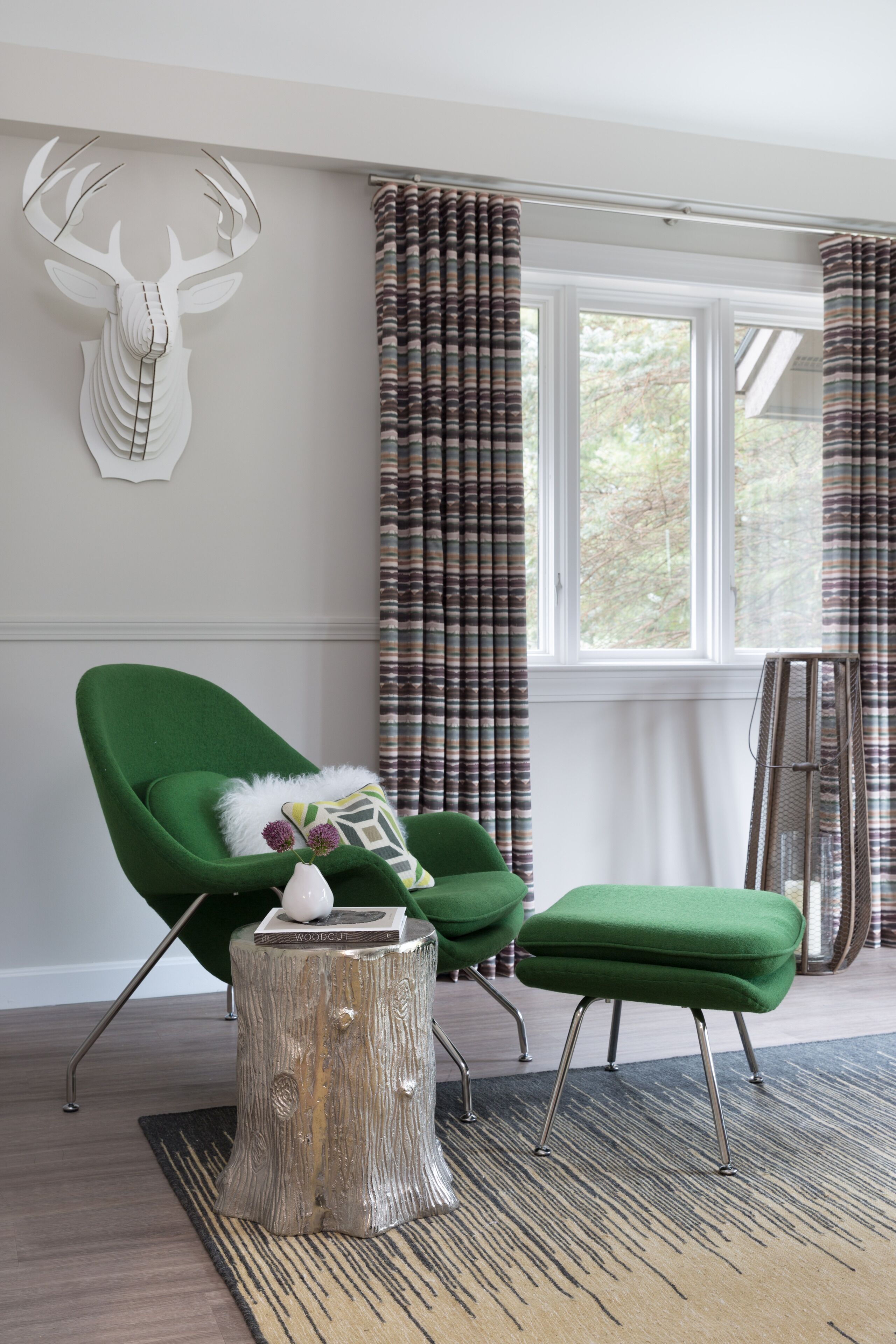 Un salon moderne avec un fauteuil vert, un pouf assorti, une petite table en bois brut et une décoration murale représentant une tête de cerf.