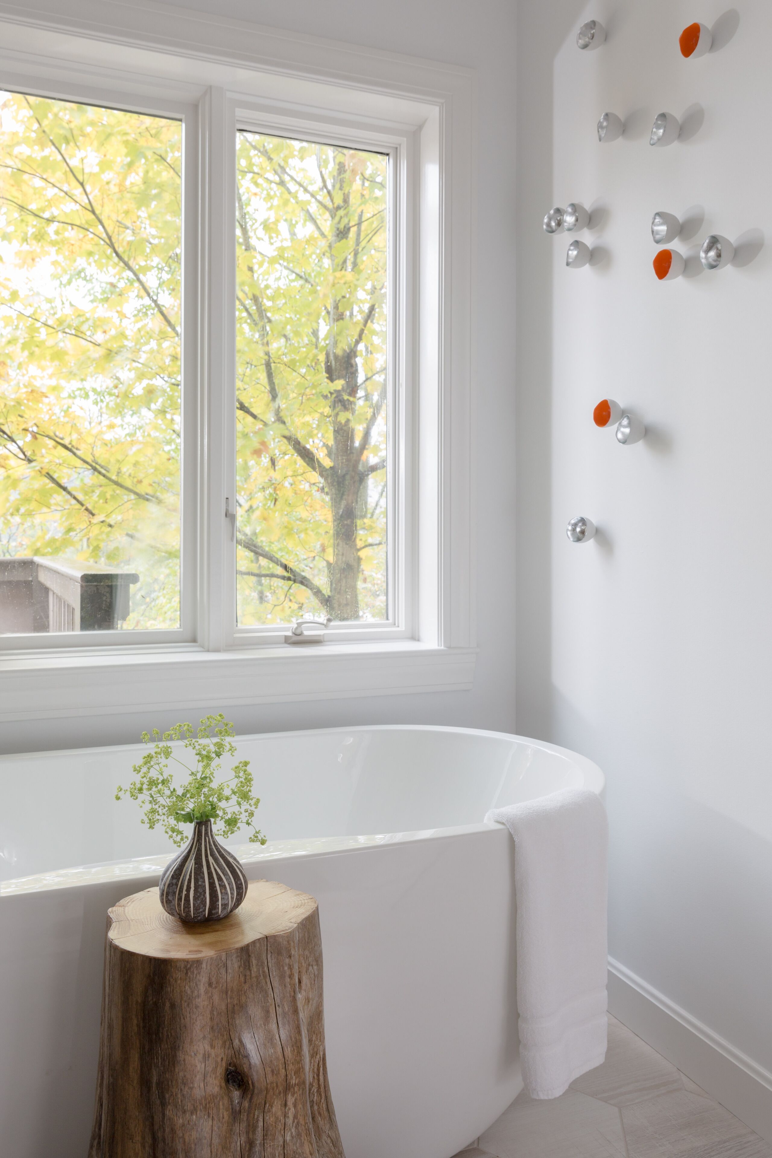 A bathroom with a white bathtub, a wooden stool with a vase, and a window with a view of trees.