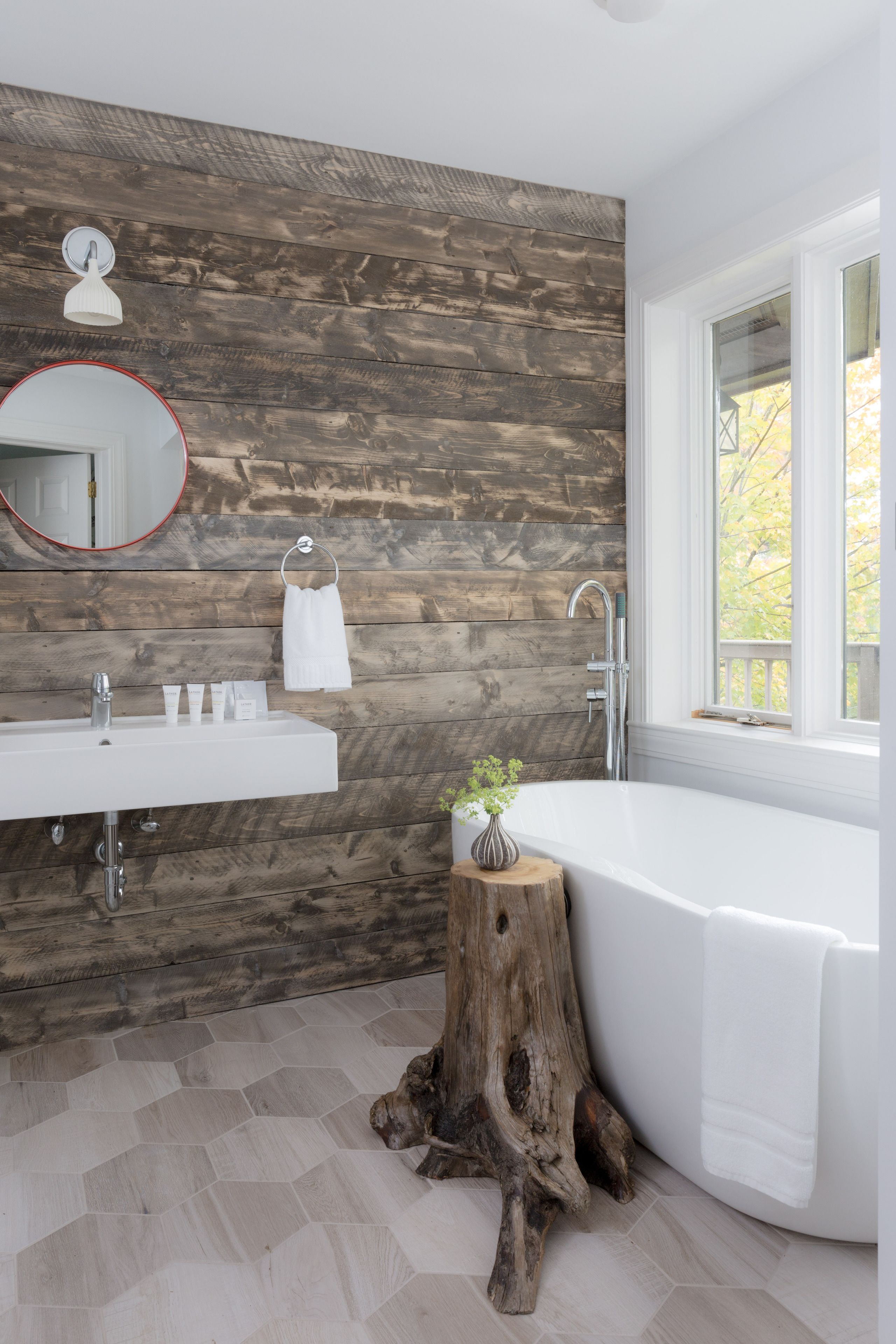 A modern bathroom with a wooden wall, a white sink, a bathtub, and a window with a view of trees.