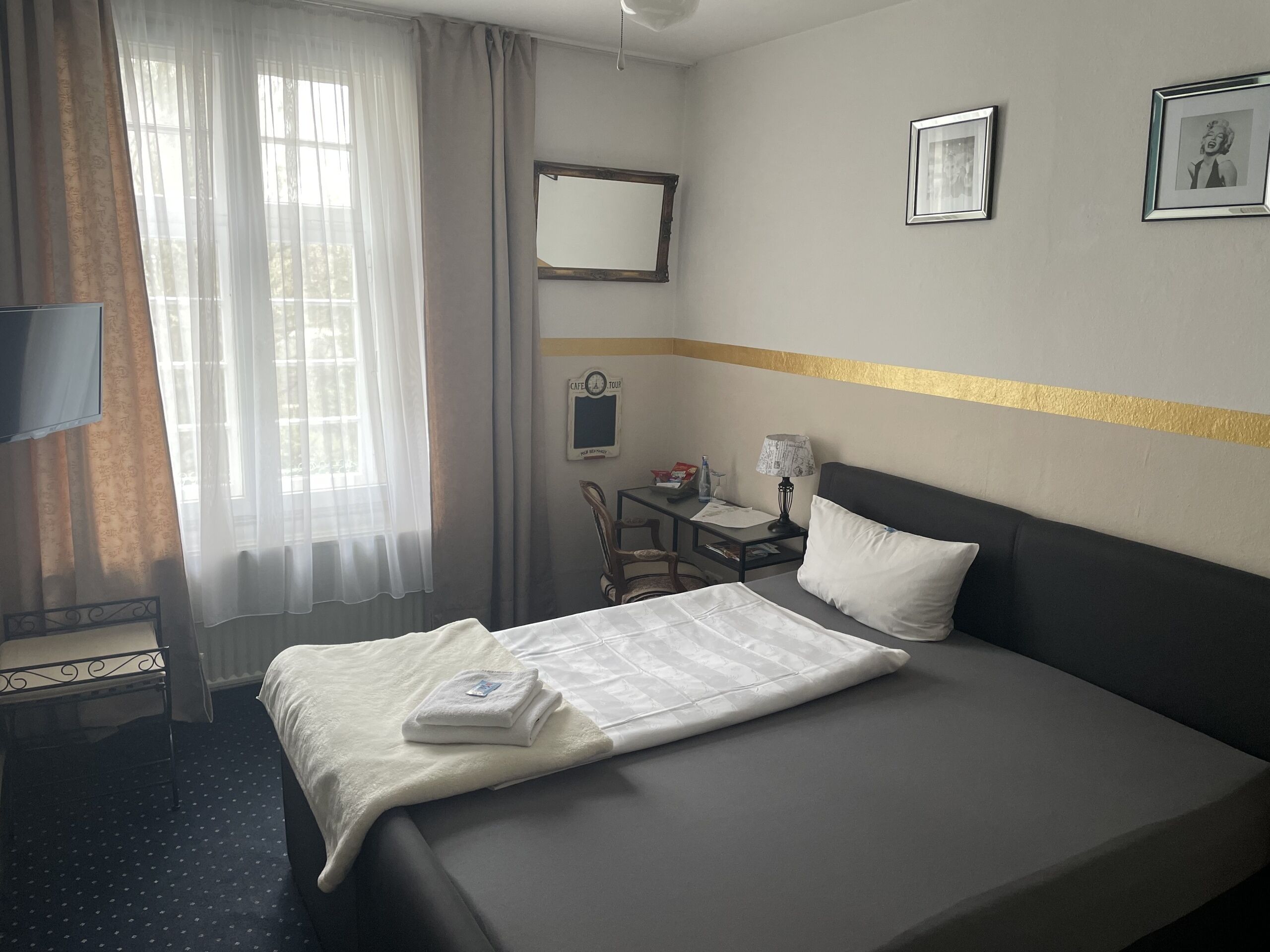Una habitación de hotel con cama, escritorio, silla, televisor y ventana con cortinas.