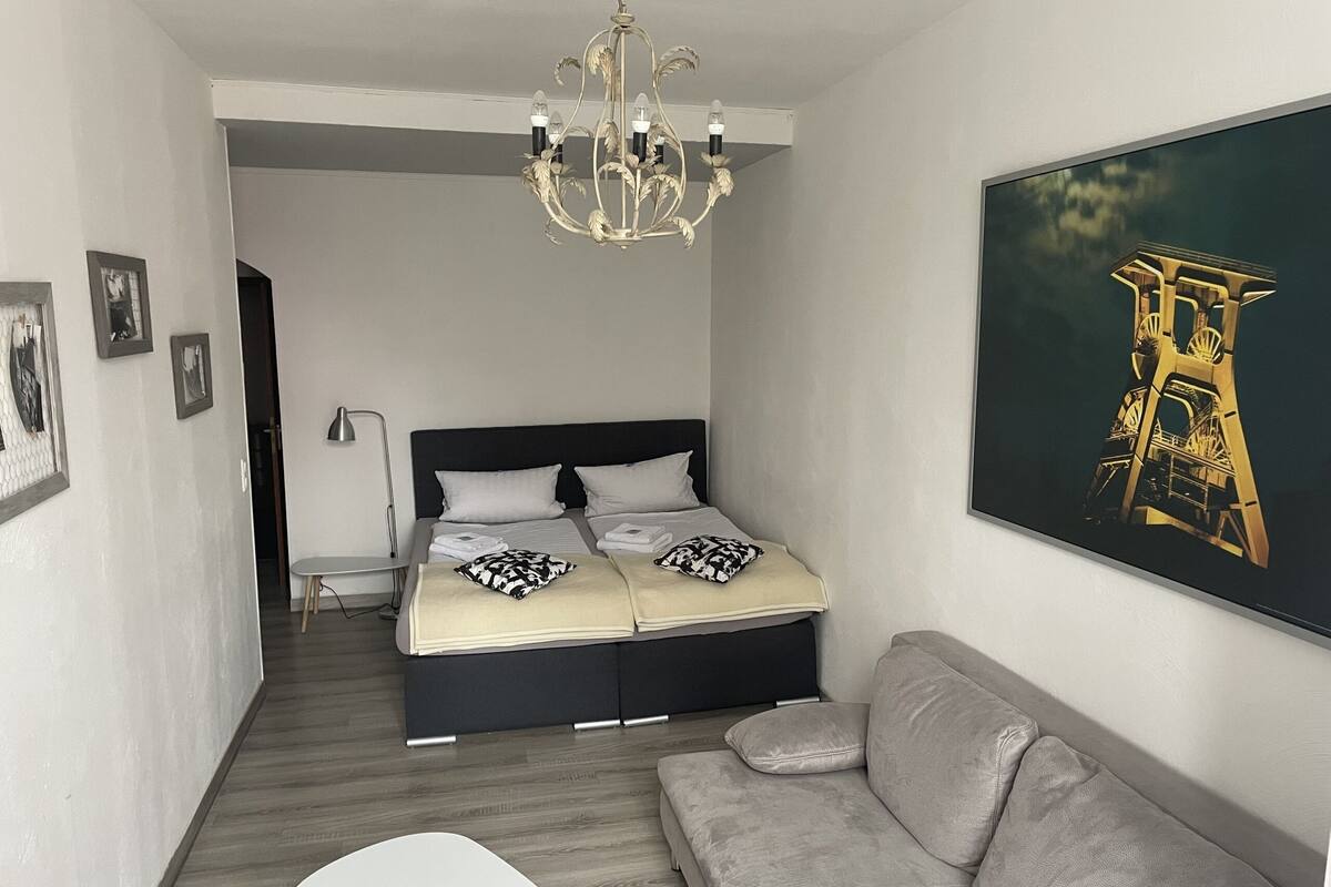 A modern bedroom with a bed, a grey sofa, a chandelier, and a painting of a tower.