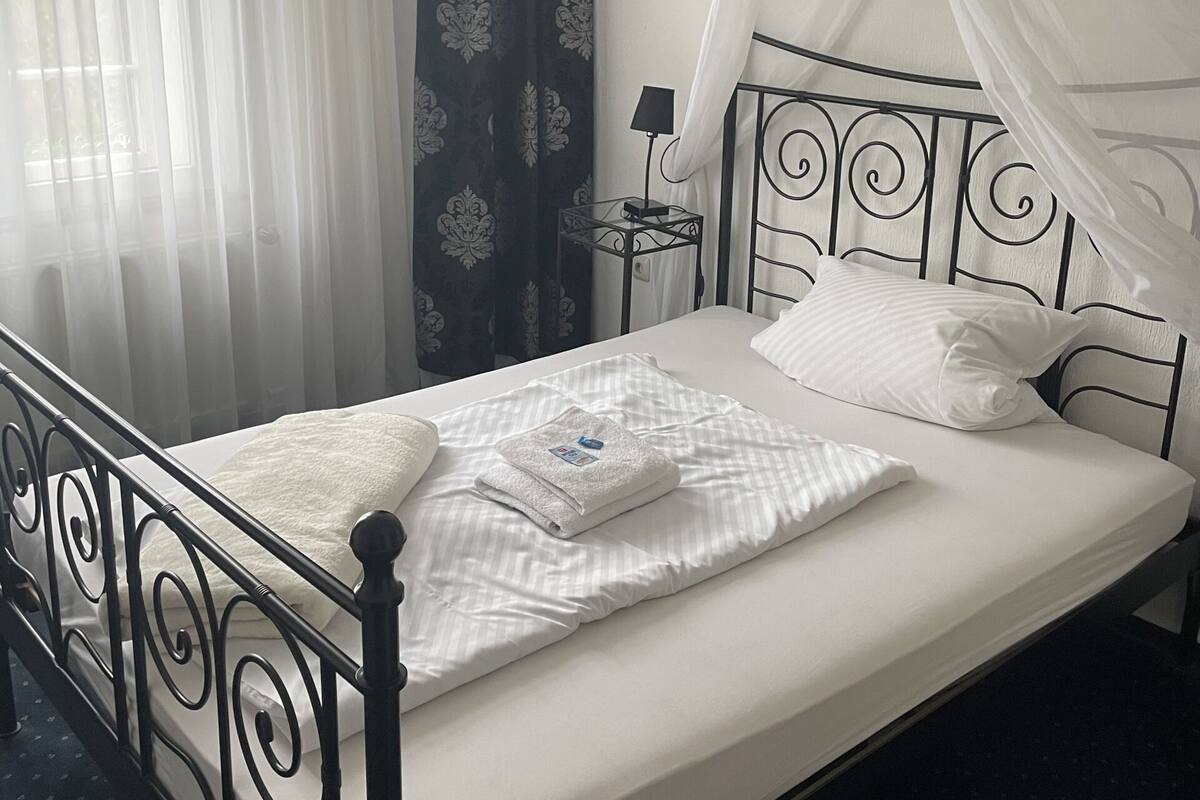A neatly made bed with white linens, a bedside table with a lamp, and a window with sheer curtains.