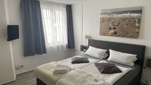 A neatly made bed with white and dark pillows, a bedside table with a lamp, and a painting of a beach scene on the wall. - Hotel Jägerhof Kettwig (Essen)