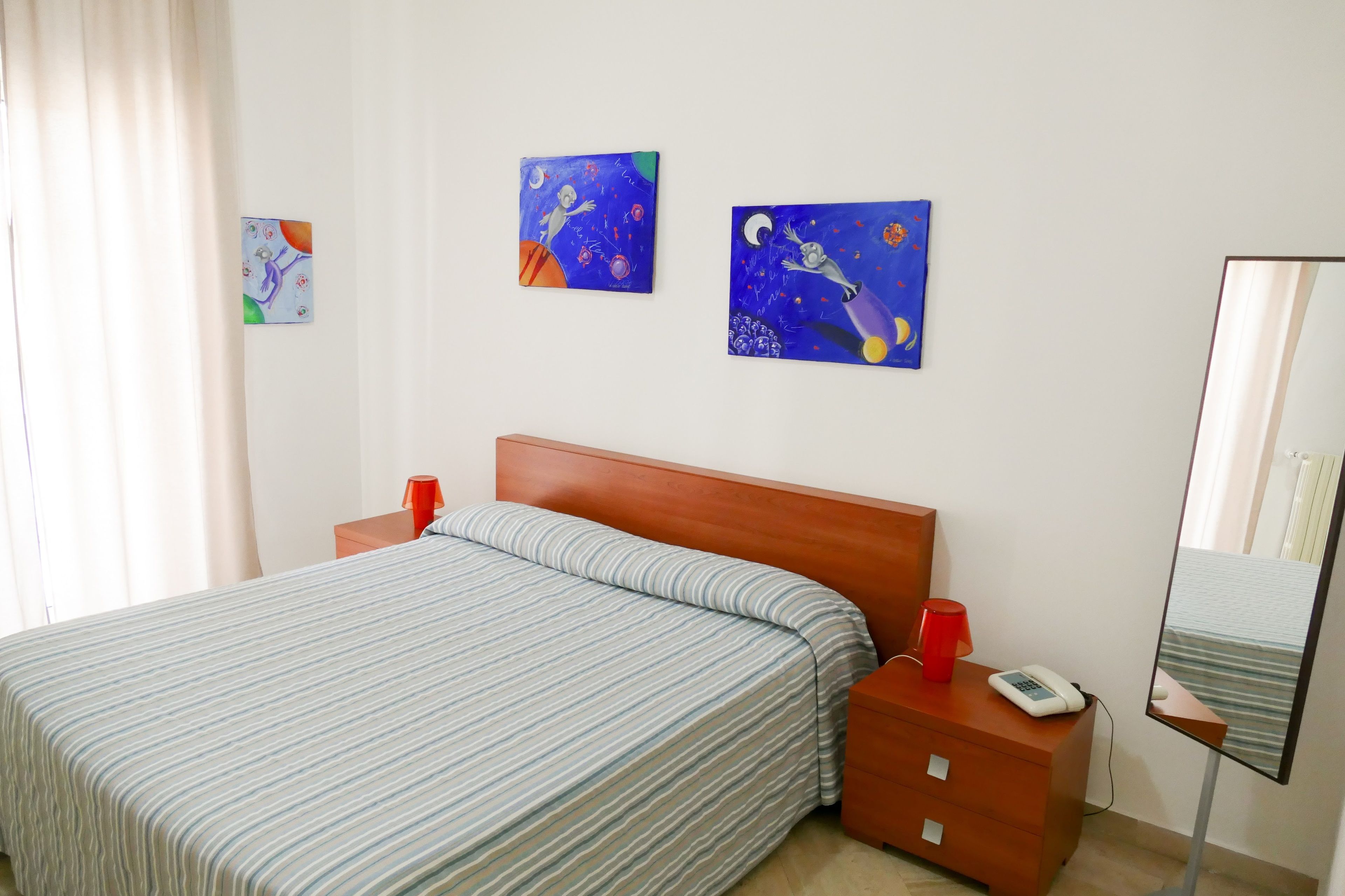 A hotel room with a bed, a nightstand, a mirror, and two paintings on the wall.