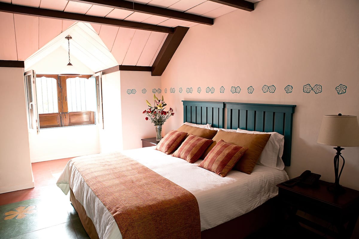 Un dormitorio con cama, mesita de noche, lámpara, un florero con flores y ventana con vistas al exterior.