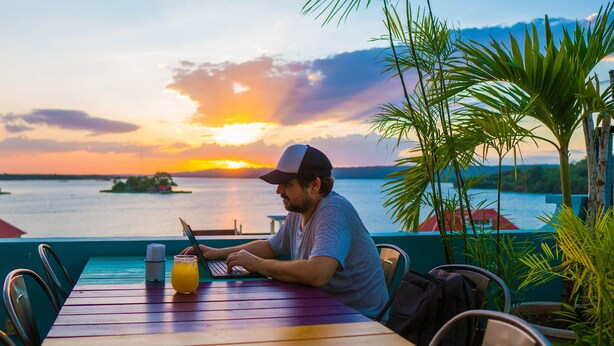 Una persona sentada a una mesa con una computadora portátil y una bebida, con vista a una masa de agua y un atardecer de fondo.