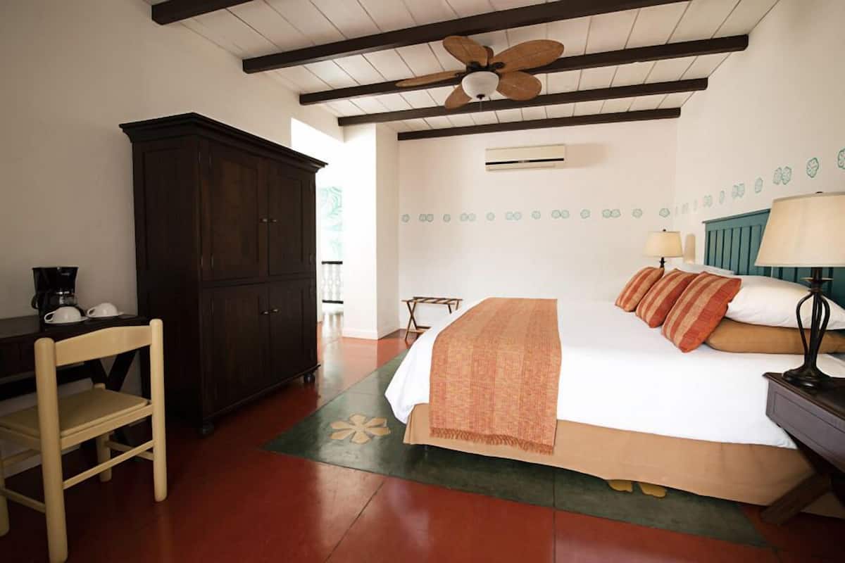 Un dormitorio con una cama, una silla de madera, un armario de madera oscura, un ventilador de techo y una alfombra verde con diseño floral.