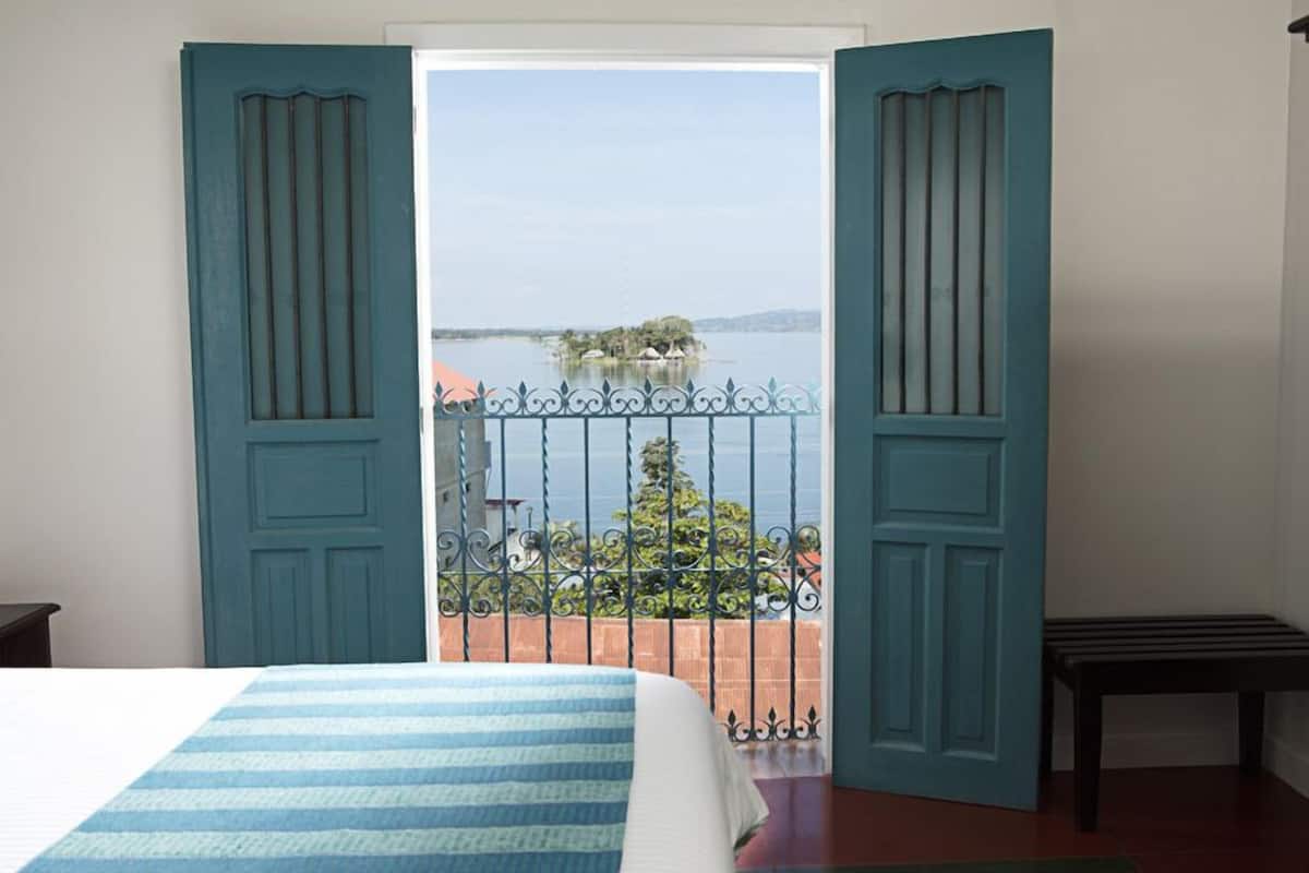 Un dormitorio con cama, un balcón que ofrece vistas a un lago y una isla, y una puerta con diseño decorativo.