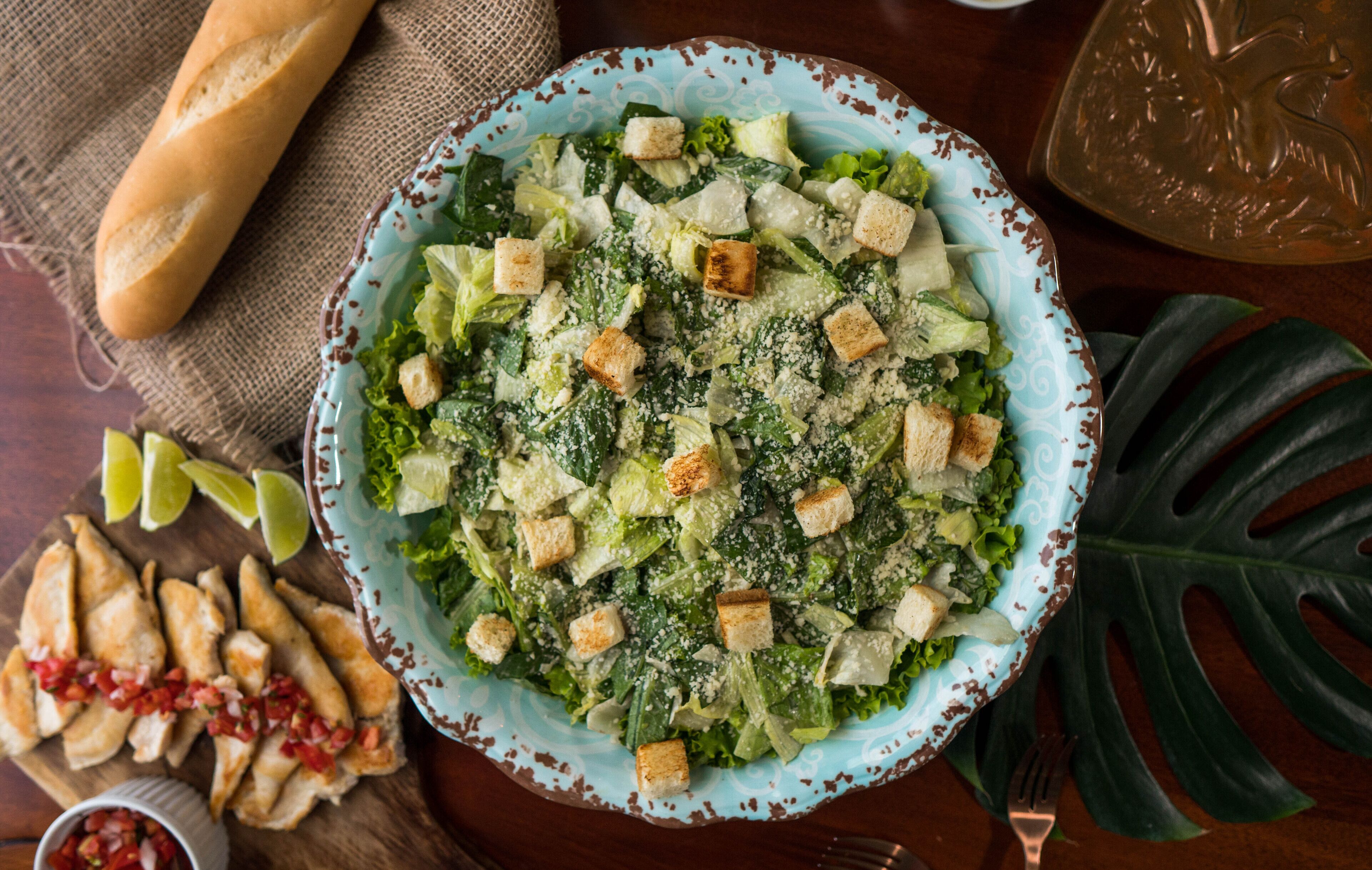 Ensalada con crutones y queso, acompañada de una baguette, gajos de lima y un cuenco pequeño de salsa.