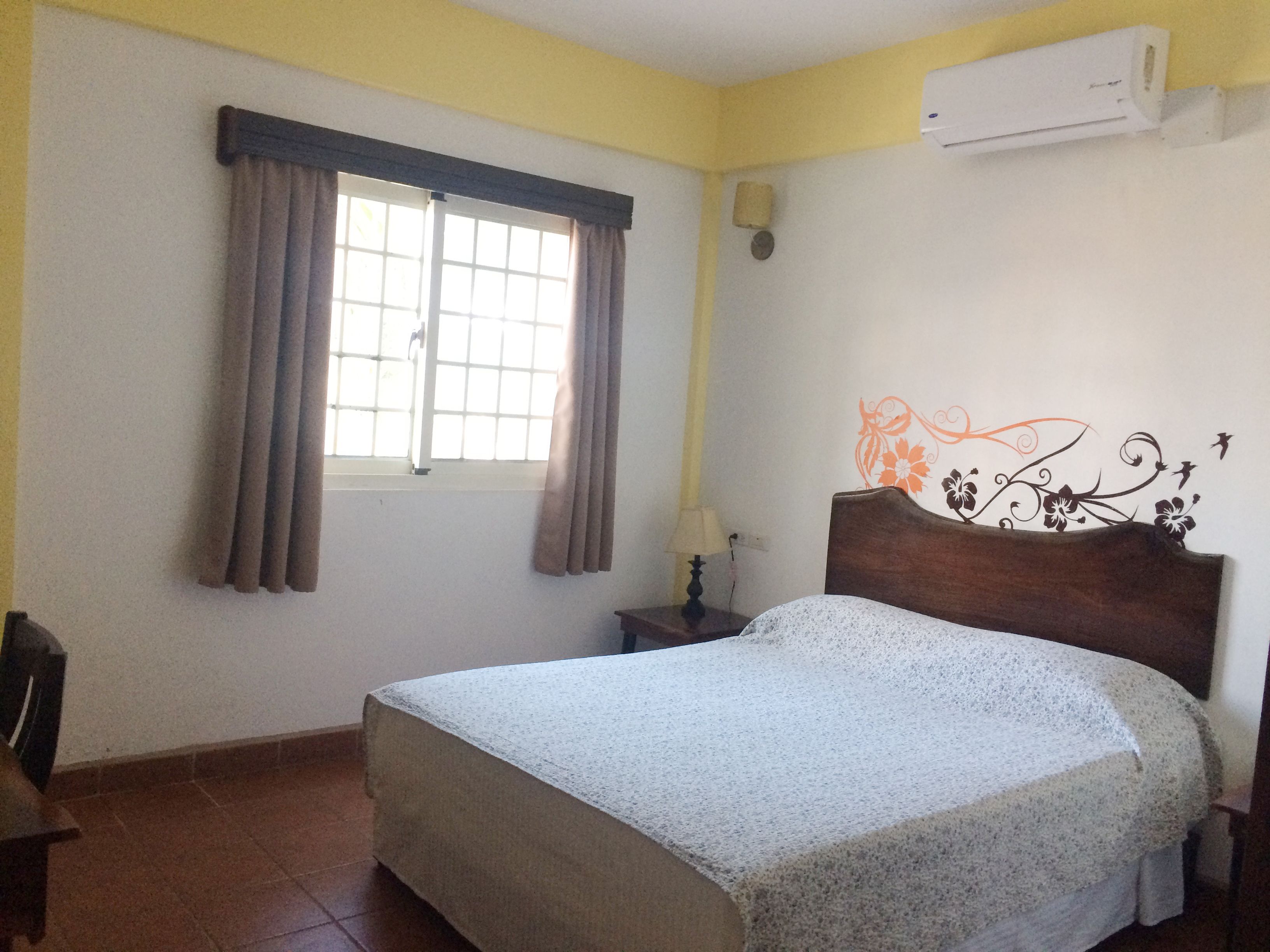A bedroom with a bed, a window with curtains, a bedside table with a lamp, and an air conditioning unit on the wall.