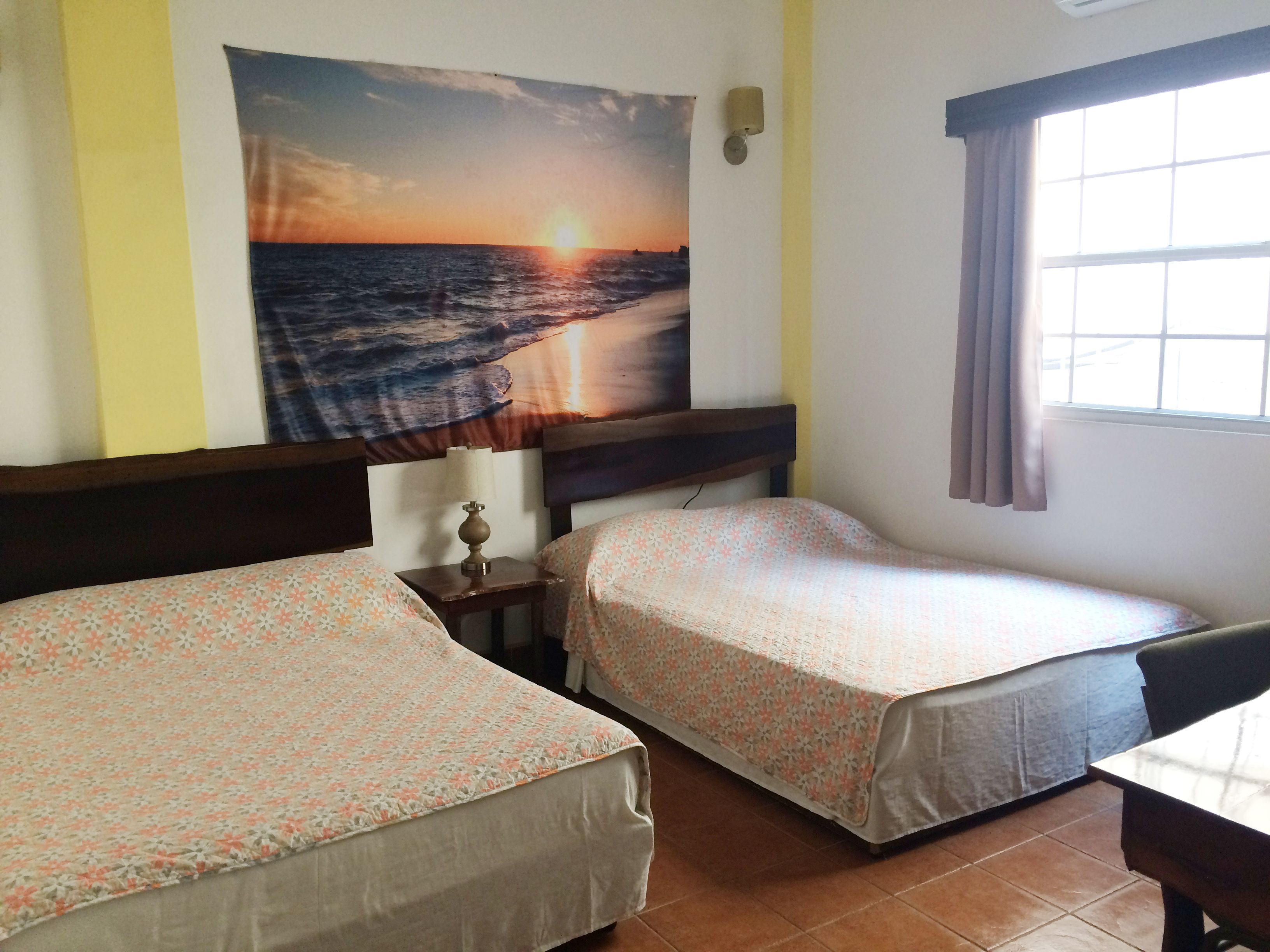 Quarto com duas camas, uma luminária na parede e uma grande pintura do pôr do sol sobre o oceano.