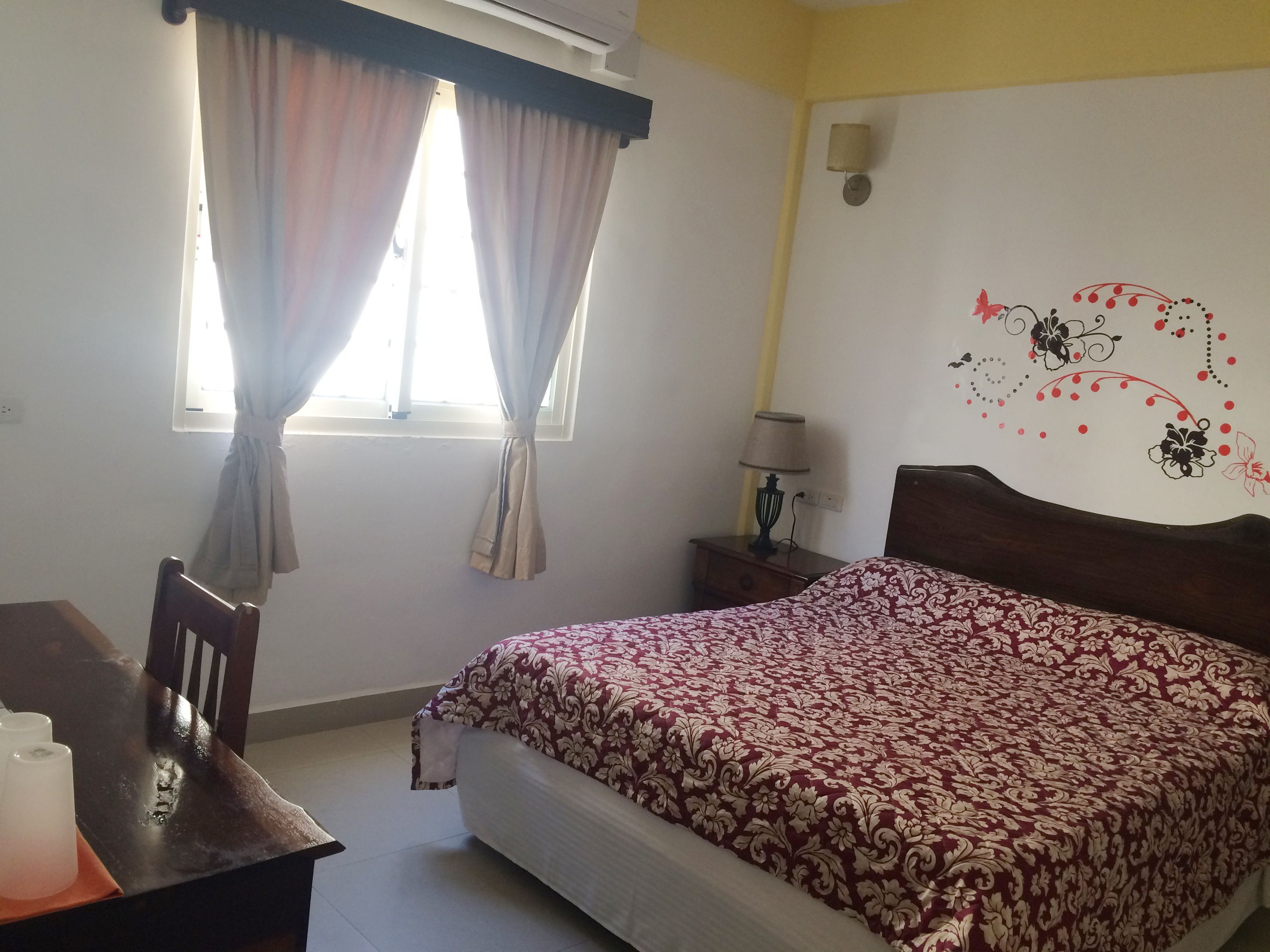 Un dormitorio con cama, mesita de noche, lámpara, silla y ventana con cortinas.