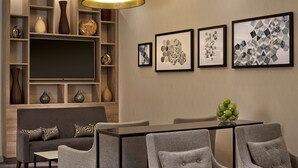 Uma sala de estar moderna com sofá, cadeiras, mesa de jantar e televisão. Há estantes com objetos decorativos e quadros nas paredes.