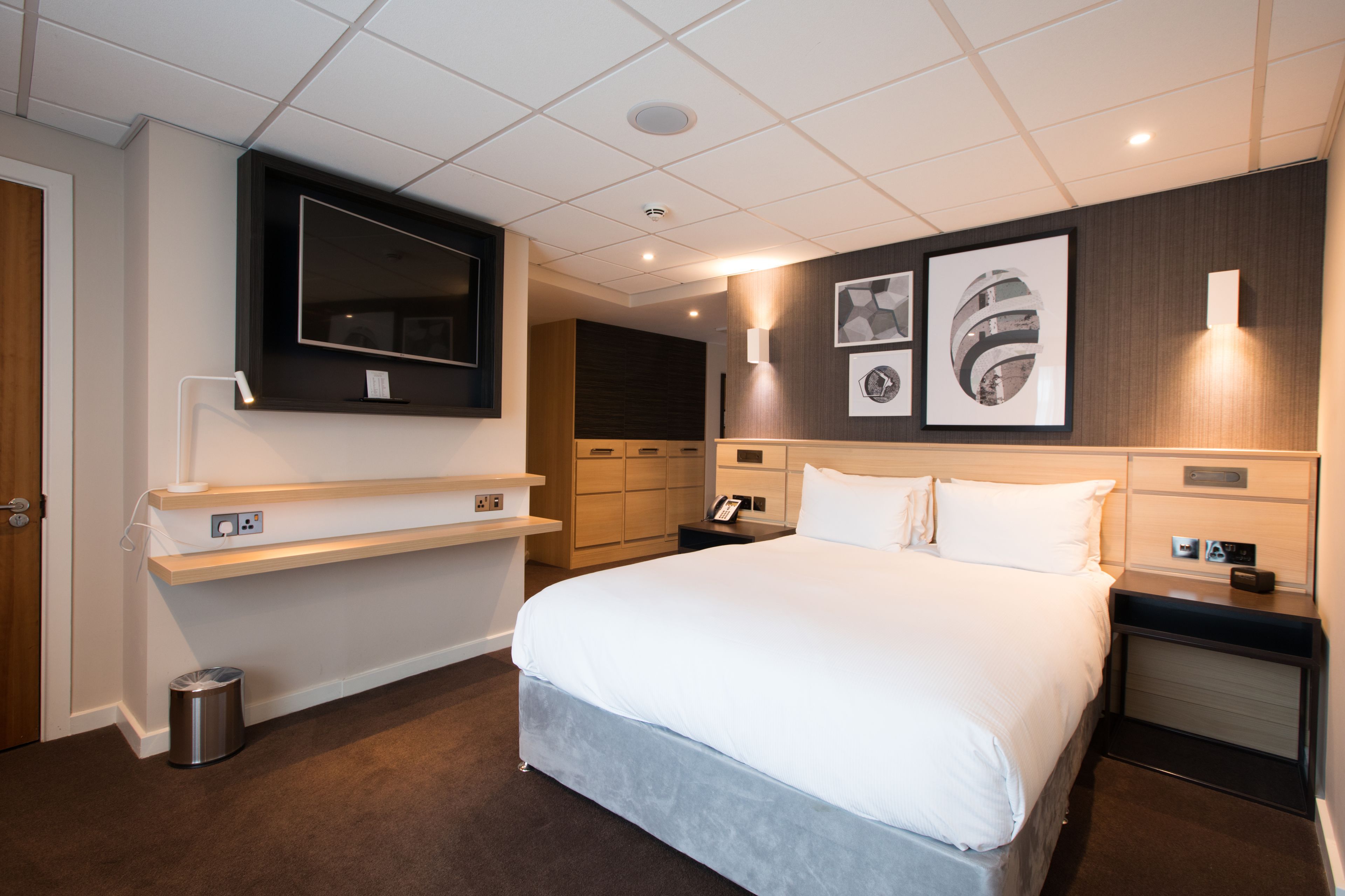 Ein modernes Hotelzimmer mit einem großen Bett, einem Wand-Fernseher, Kunstwerken an der Wand und einem kleinen Schreibtisch mit Telefon.