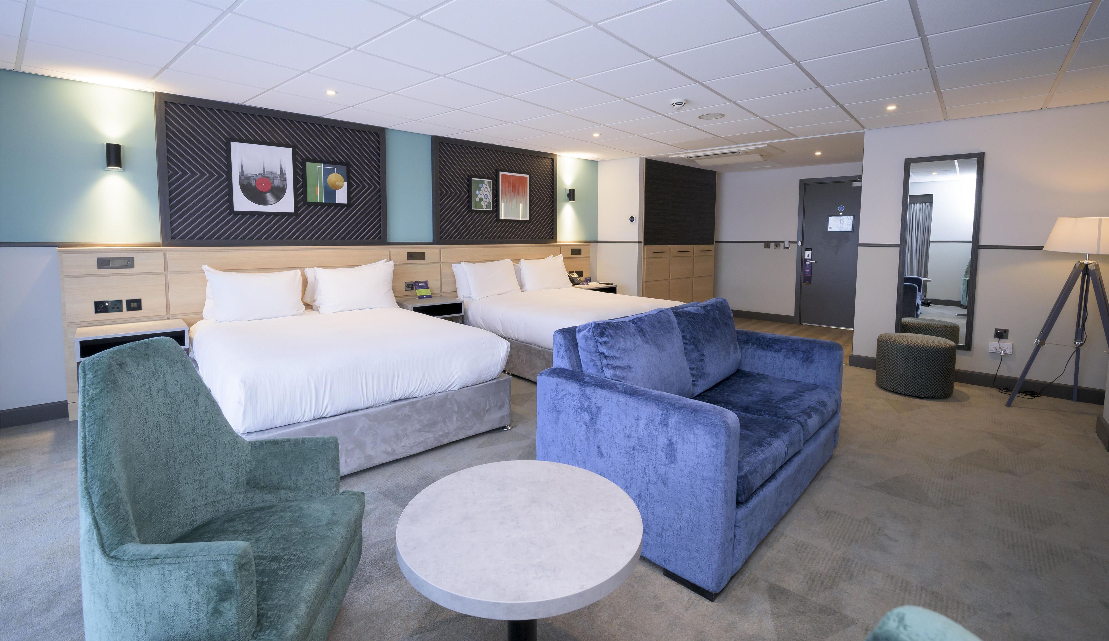 Une chambre d’hôtel avec deux lits, une table ronde, un canapé bleu et un fauteuil vert.