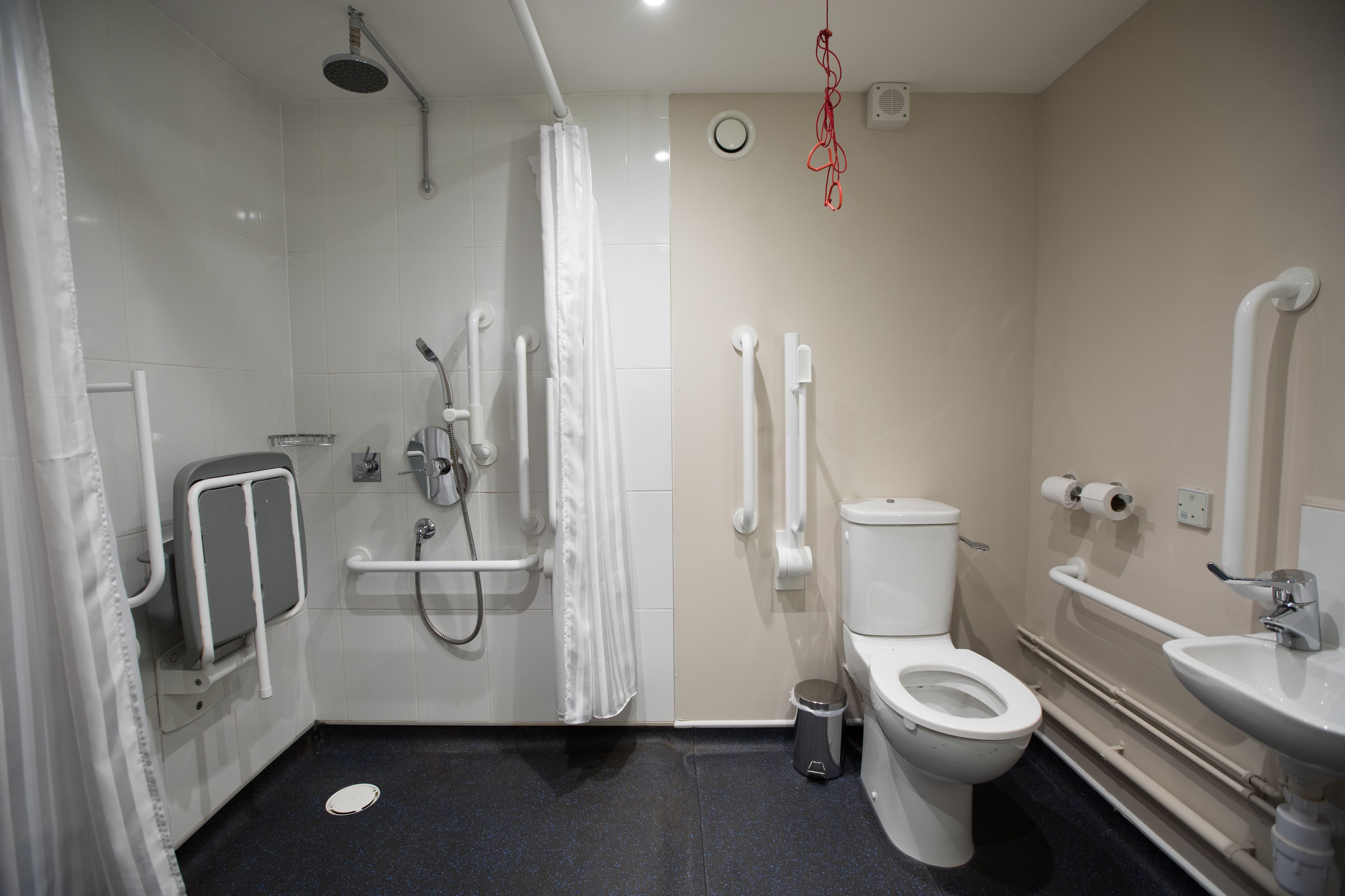 Une salle de bain moderne équipée d’une douche, de toilettes et d’un lavabo, et aménagée pour faciliter l’accès aux personnes à mobilité réduite, avec notamment des barres d’appui et un siège de douche accessible aux fauteuils roulants.