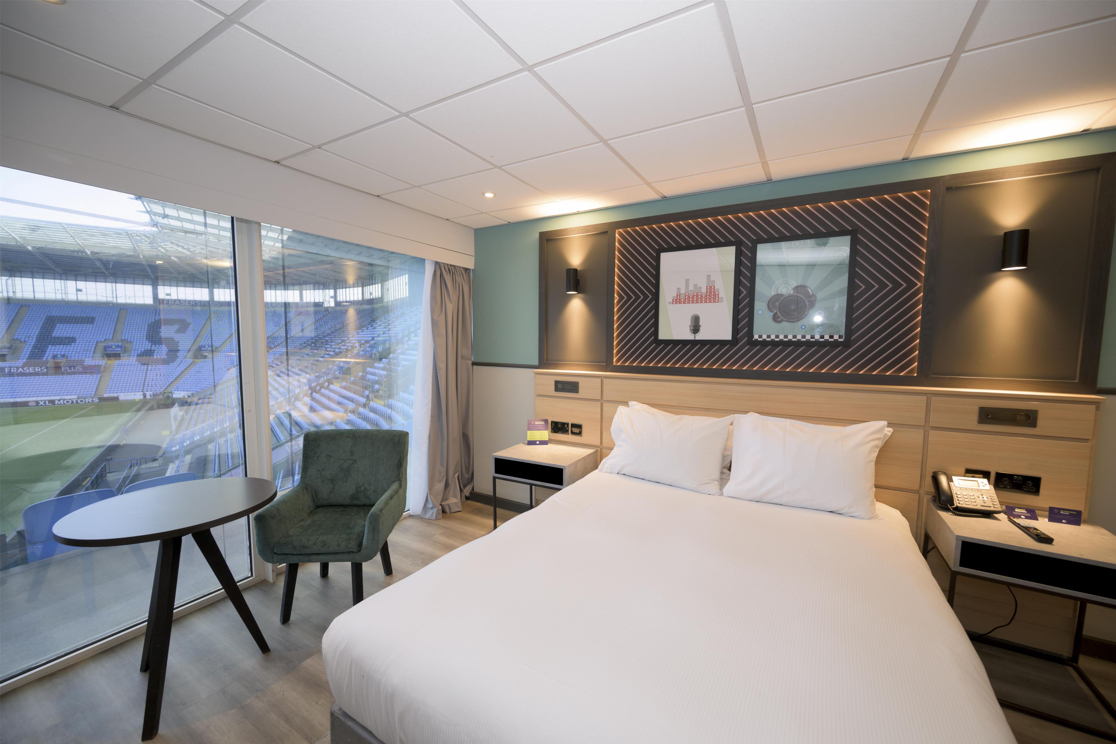 Une chambre d’hôtel comprenant un lit, un bureau, une chaise, une grande fenêtre donnant sur un stade et une œuvre d’art accrochée au mur.