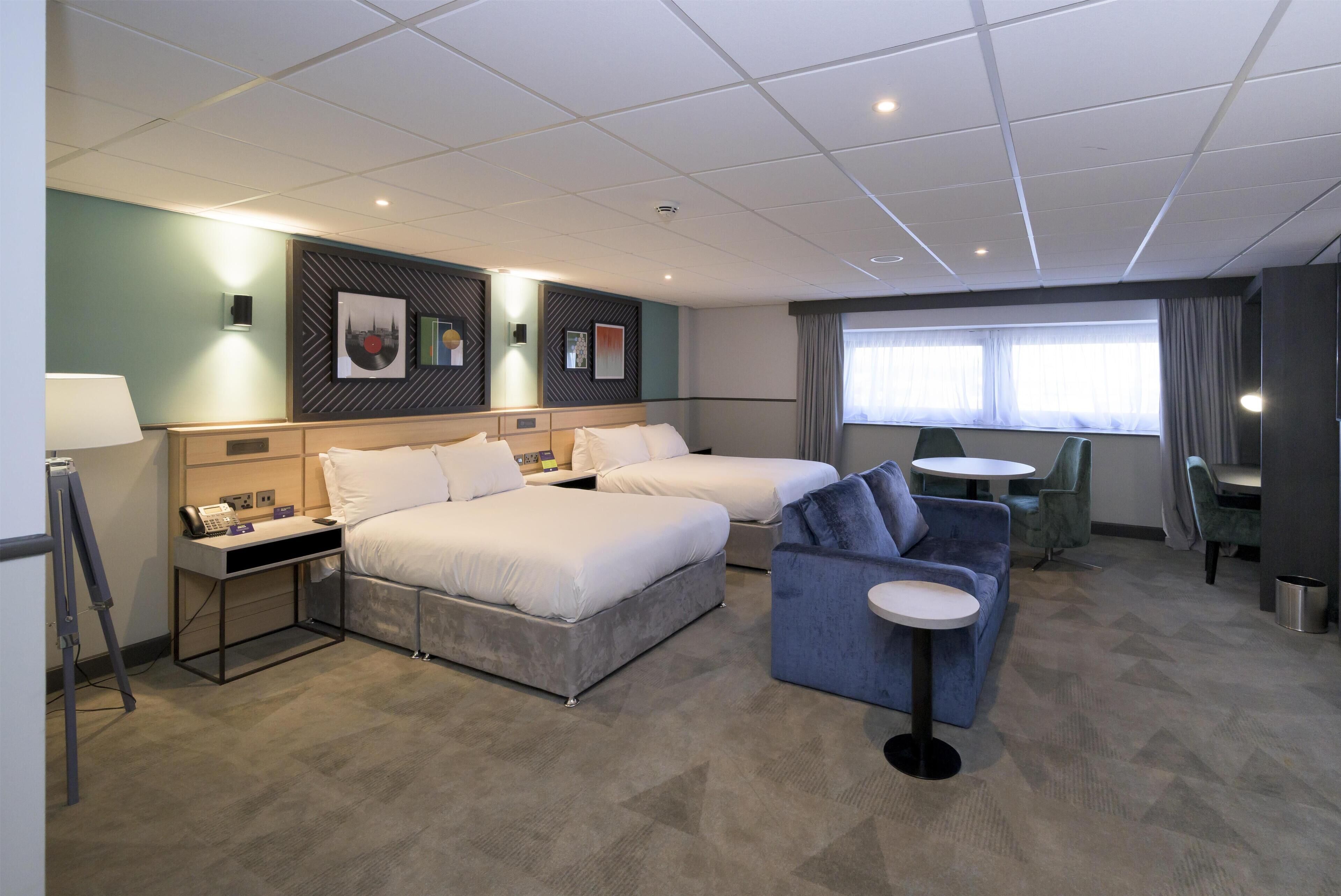 A hotel room with two beds, a desk, a small sofa, a round table, and a window with curtains.