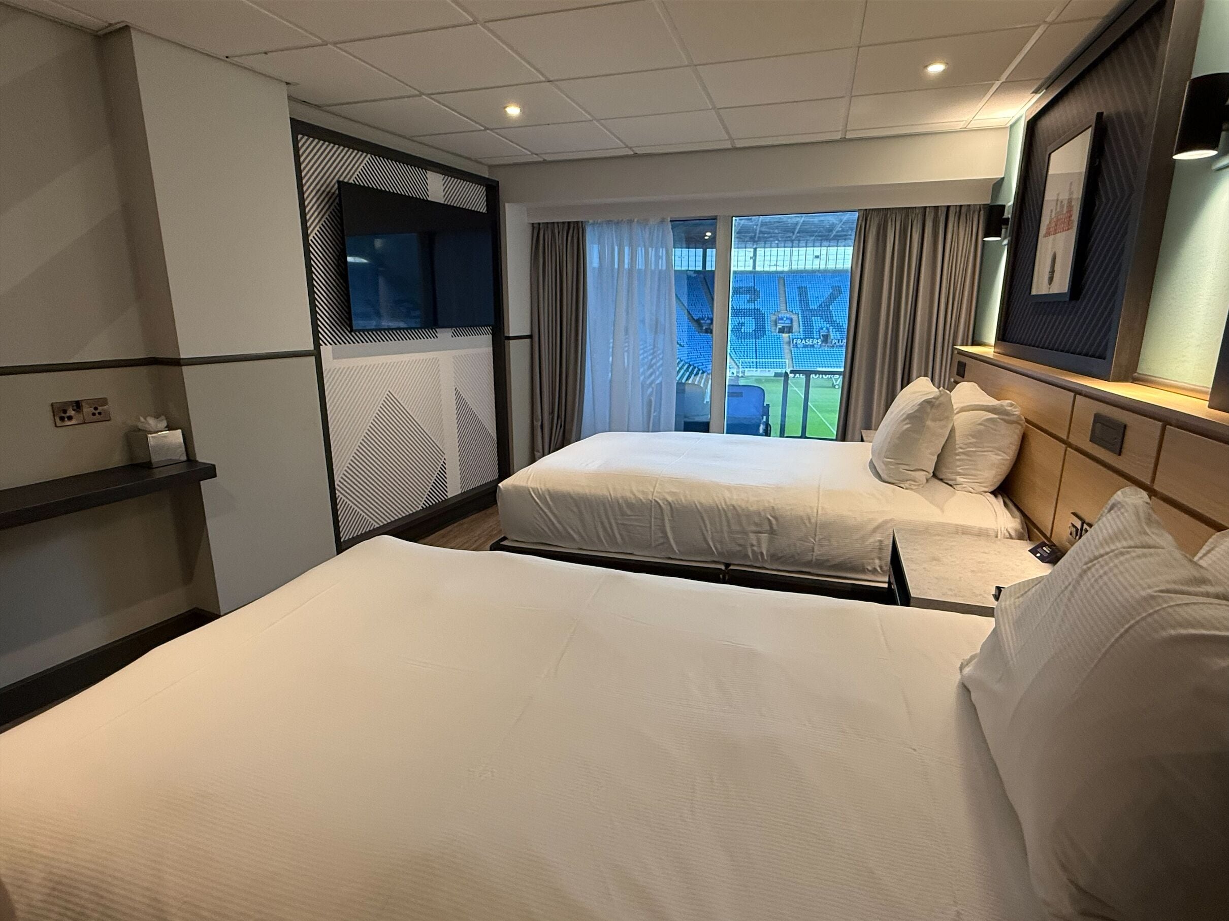 A hotel room with two beds, a TV, a window with a view of a stadium, and a shelf with a tissue box.