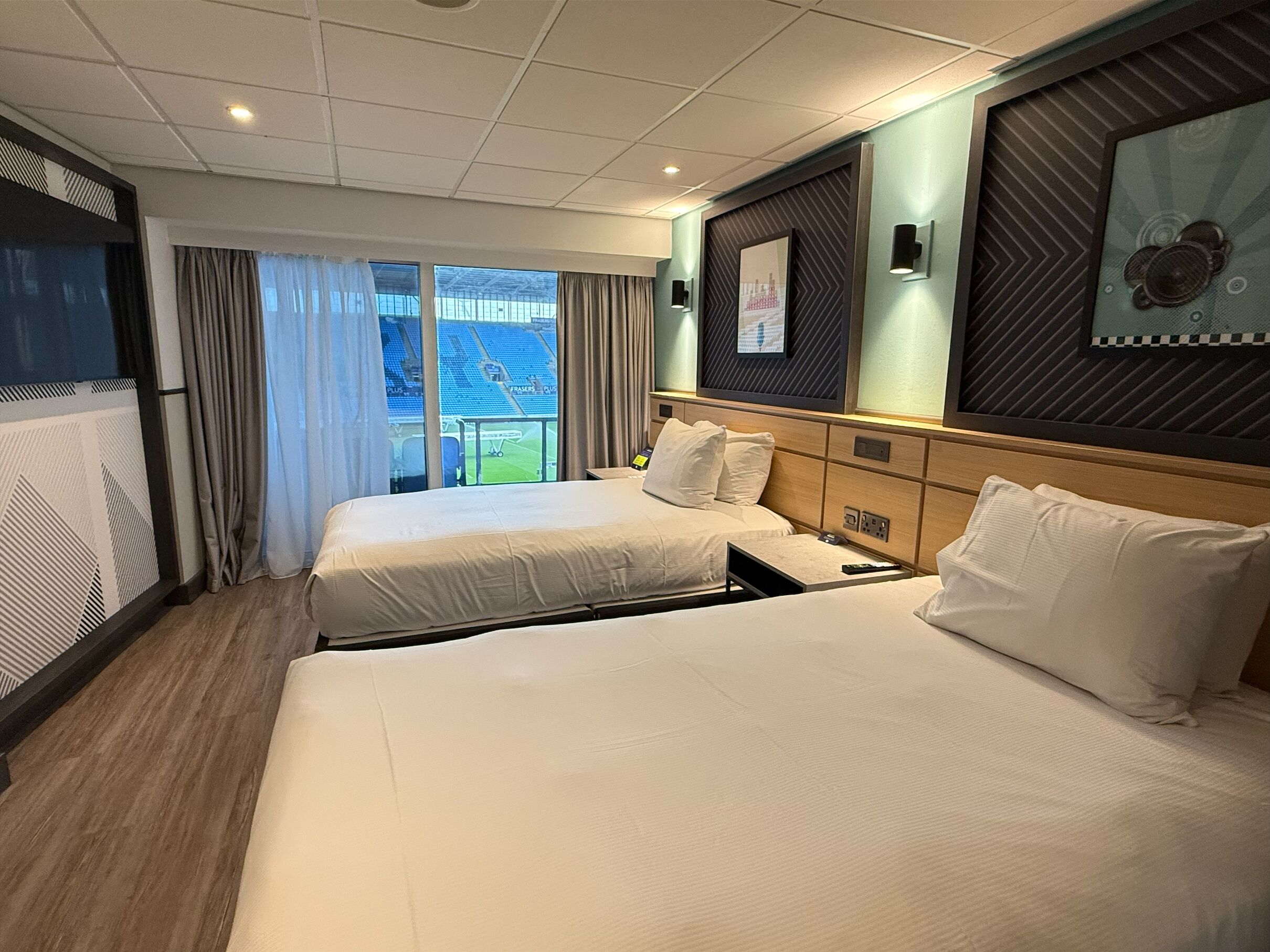 Habitación de hotel con dos camas, un ventanal grande con cortinas, un televisor de pantalla plana y una obra de arte colgada en la pared.