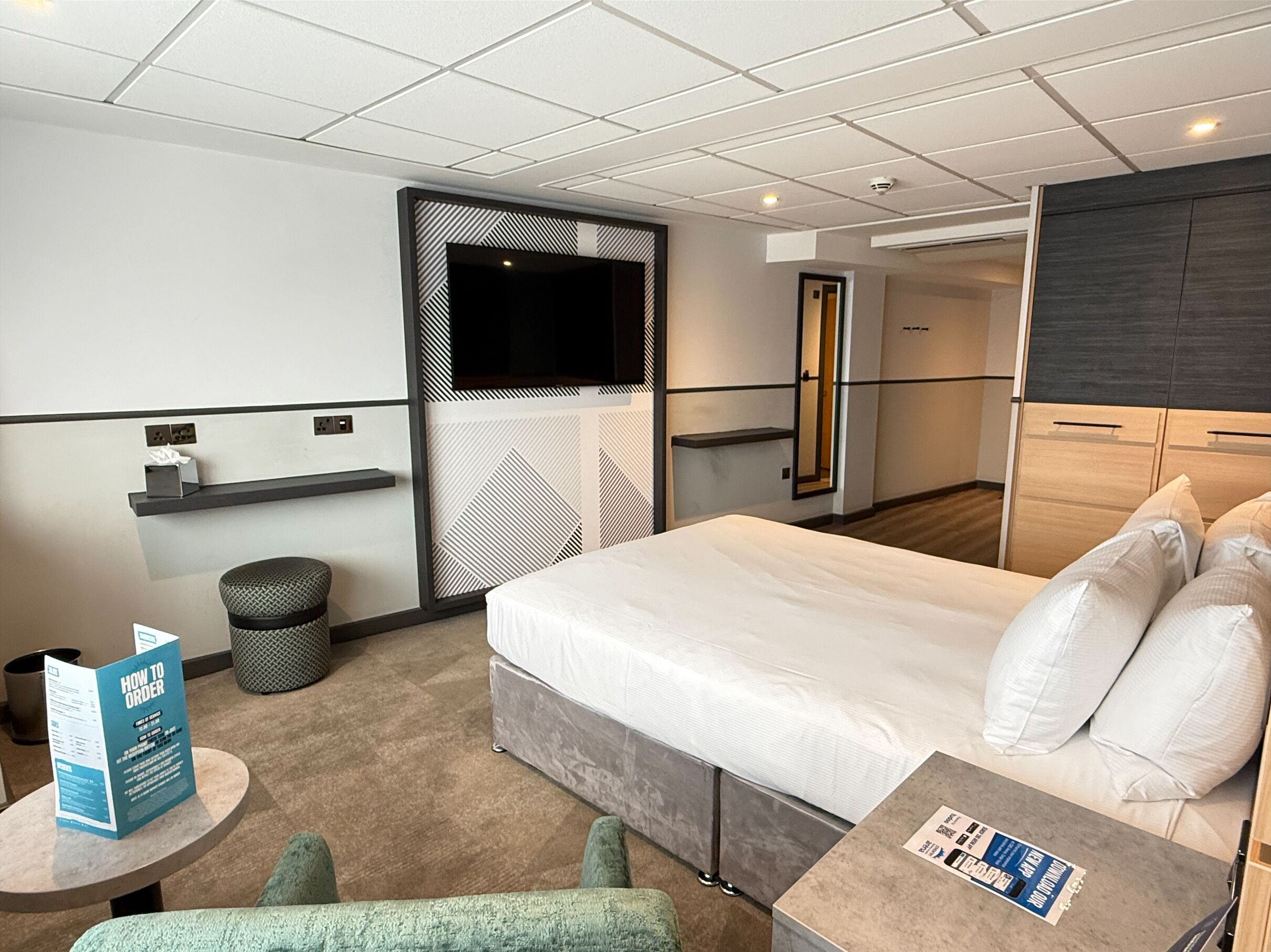 Una habitación de hotel moderna con una cama grande, un televisor montado en la pared, una pequeña mesa redonda y un rincón de descanso con una silla.