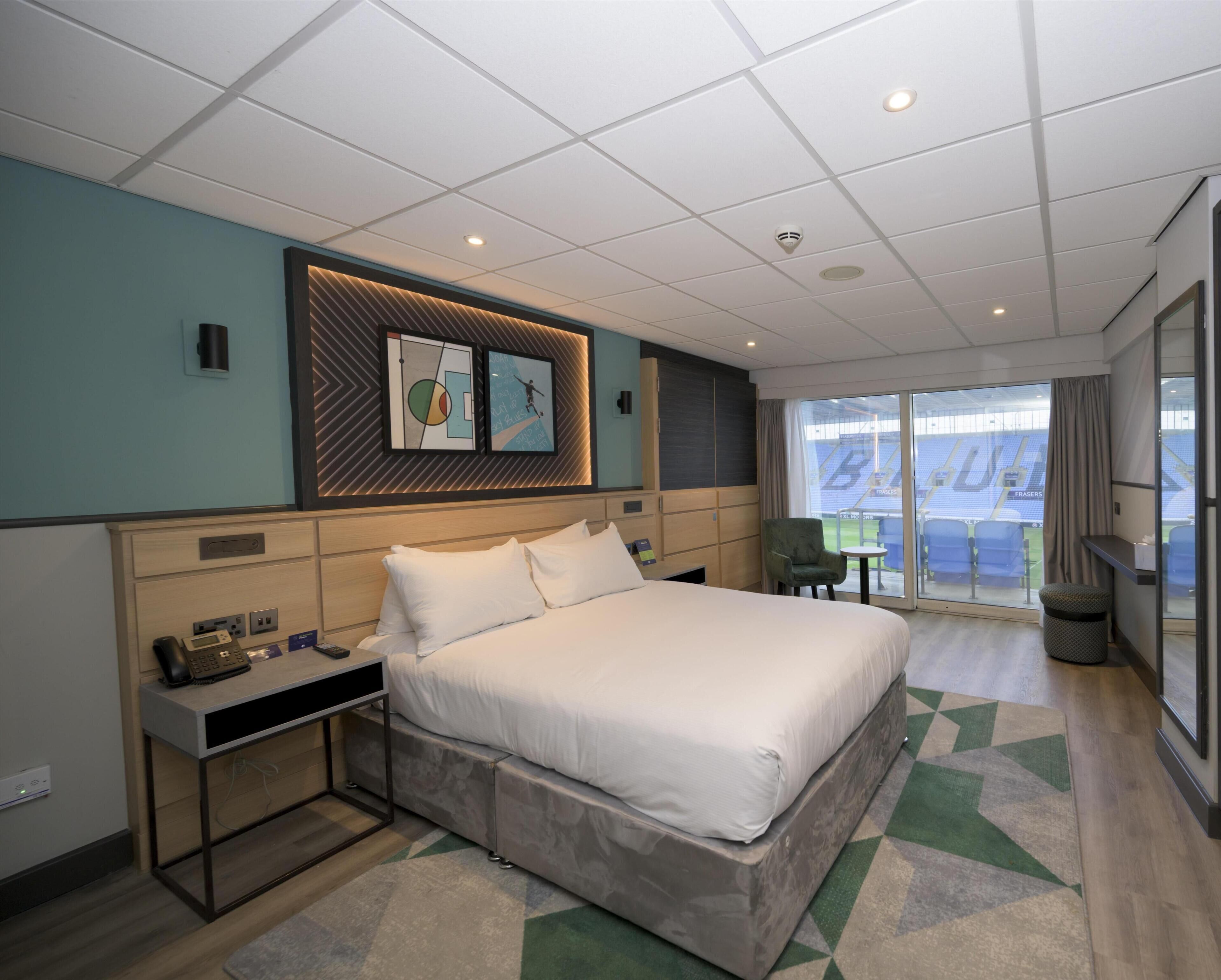 A modern hotel room with a large bed, a desk, a chair, and a view of a stadium through the glass door.