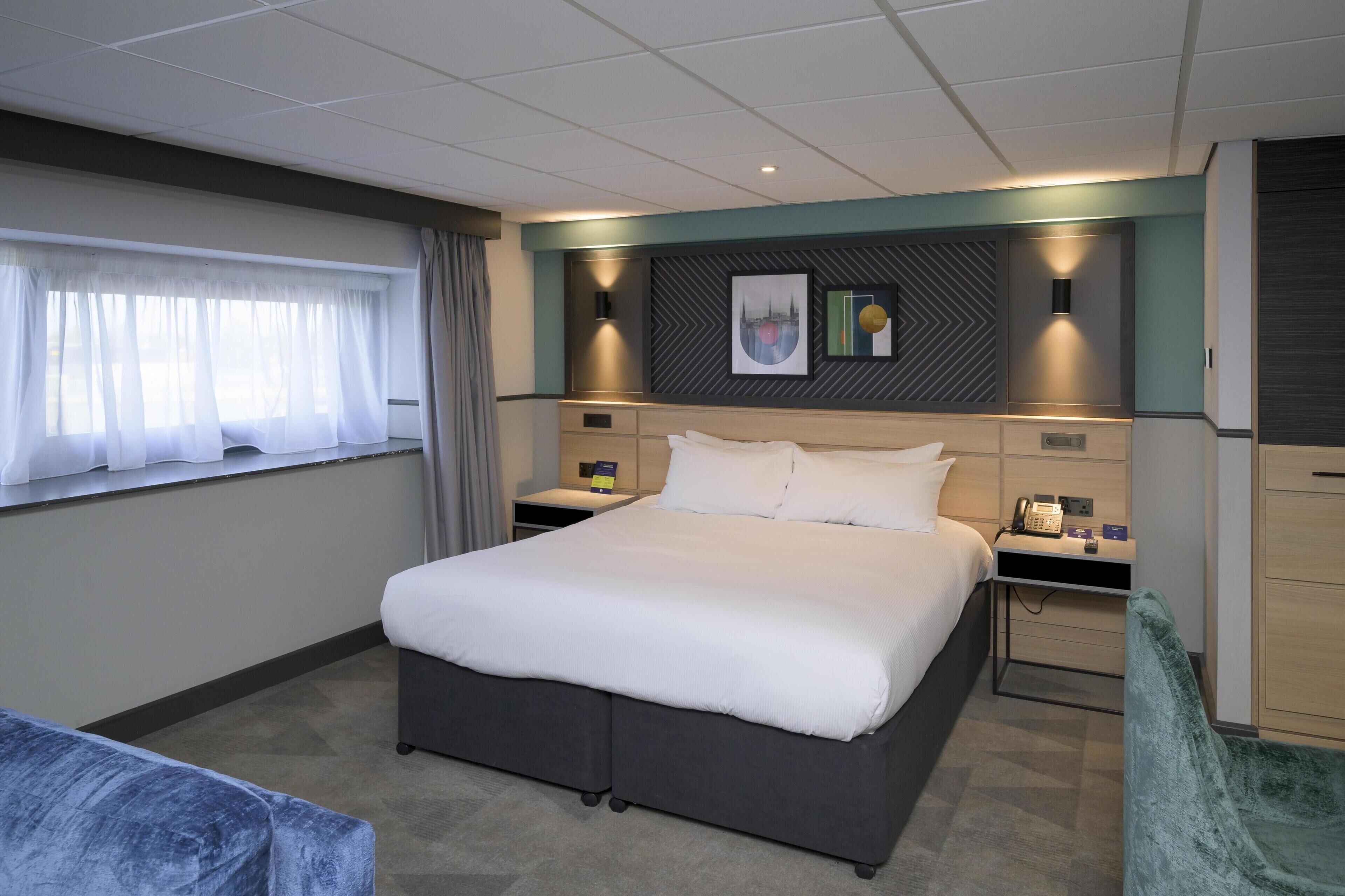 Une chambre d’hôtel avec un grand lit, des tables de chevet, un bureau, une chaise et une fenêtre avec des rideaux.