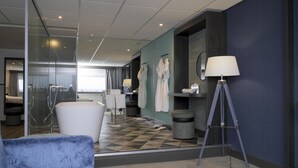 Quarto de hotel moderno com uma banheira grande, uma área de estar com uma poltrona azul, um abajur e um espelho refletindo o quarto.