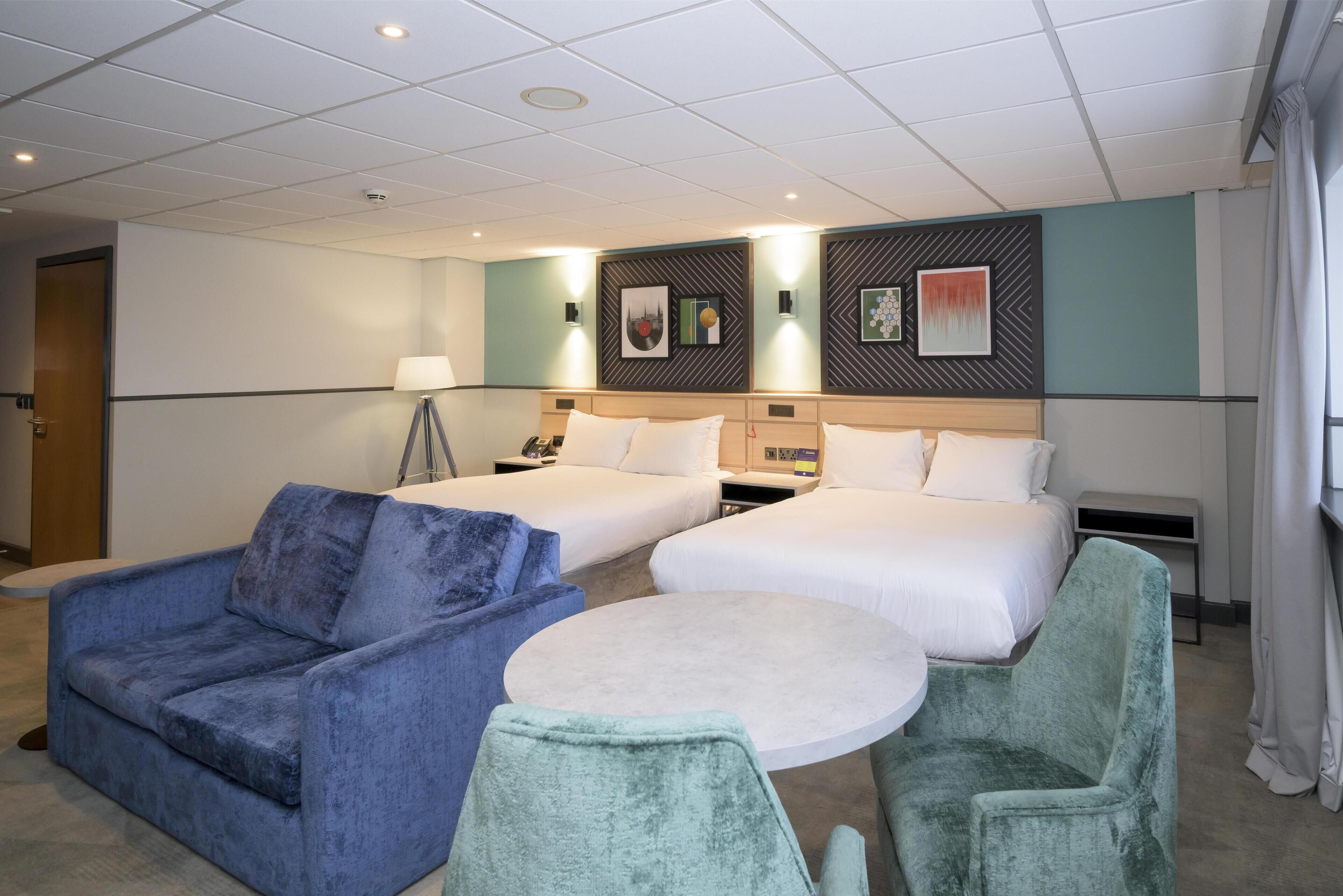 A hotel room with two beds, a sofa, a round table, and a lamp.