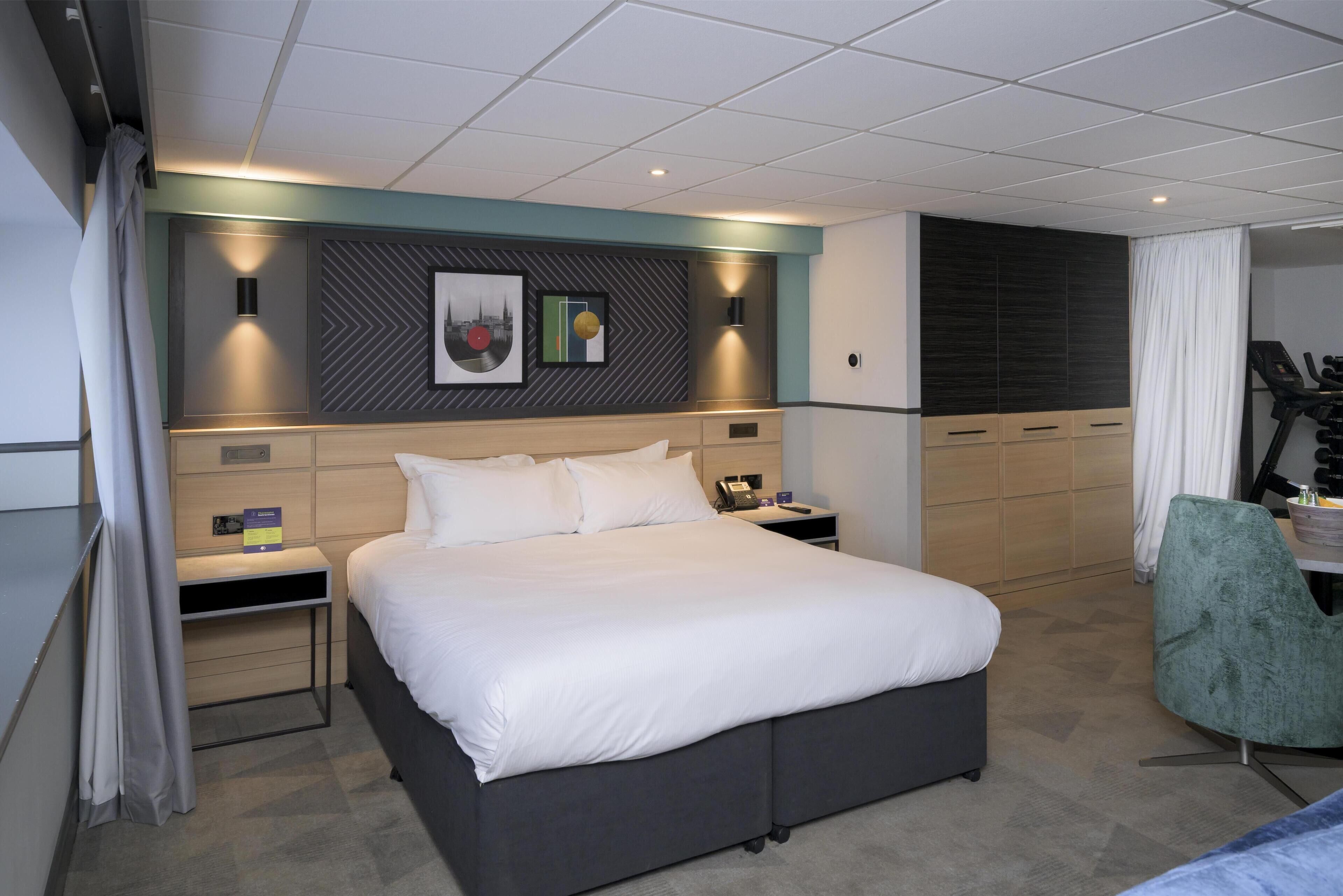 A hotel room with a large bed, bedside tables, a desk with a chair, a treadmill, and a wall with a geometric pattern and artwork.
