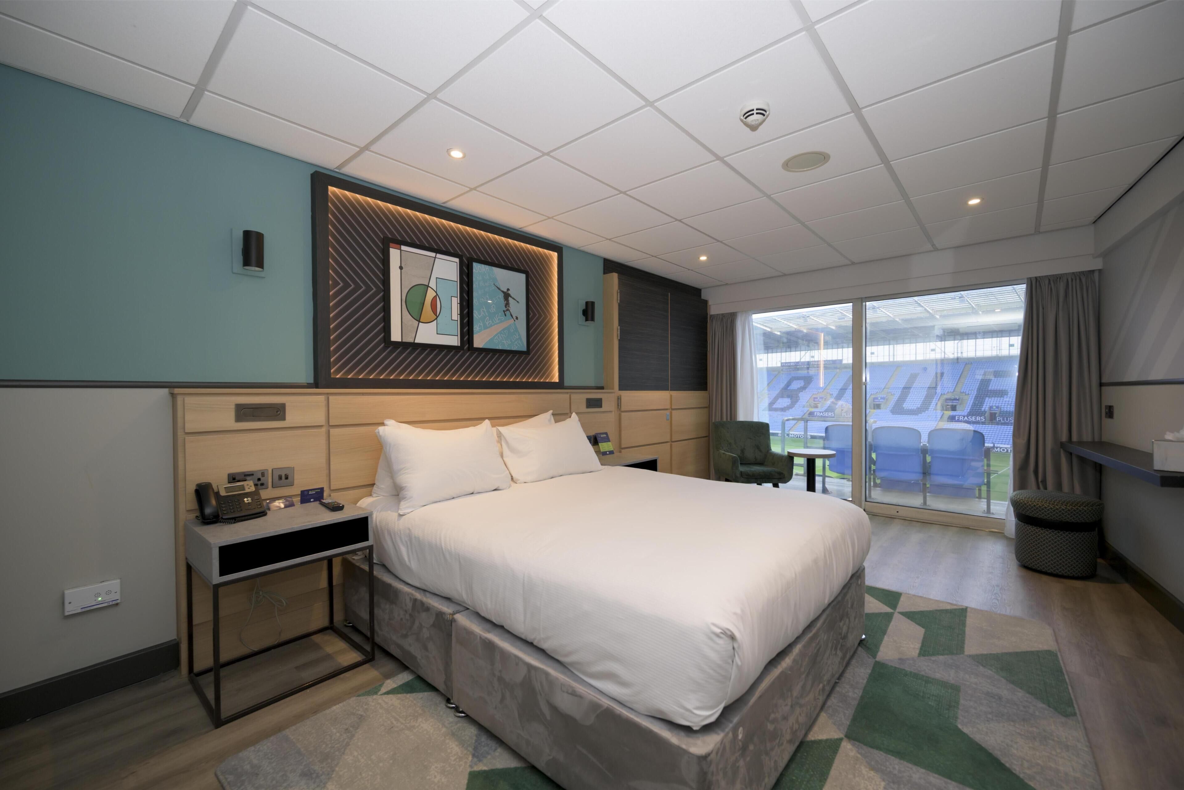 A modern hotel room with a large bed, bedside tables, a desk, and a view of a stadium.