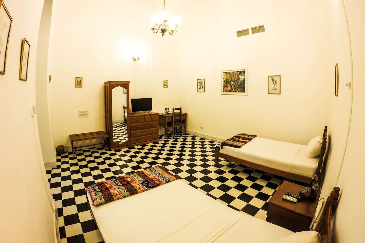A room with a checkered floor, a bed, a desk, a chair, a dresser, a mirror, and a chandelier.